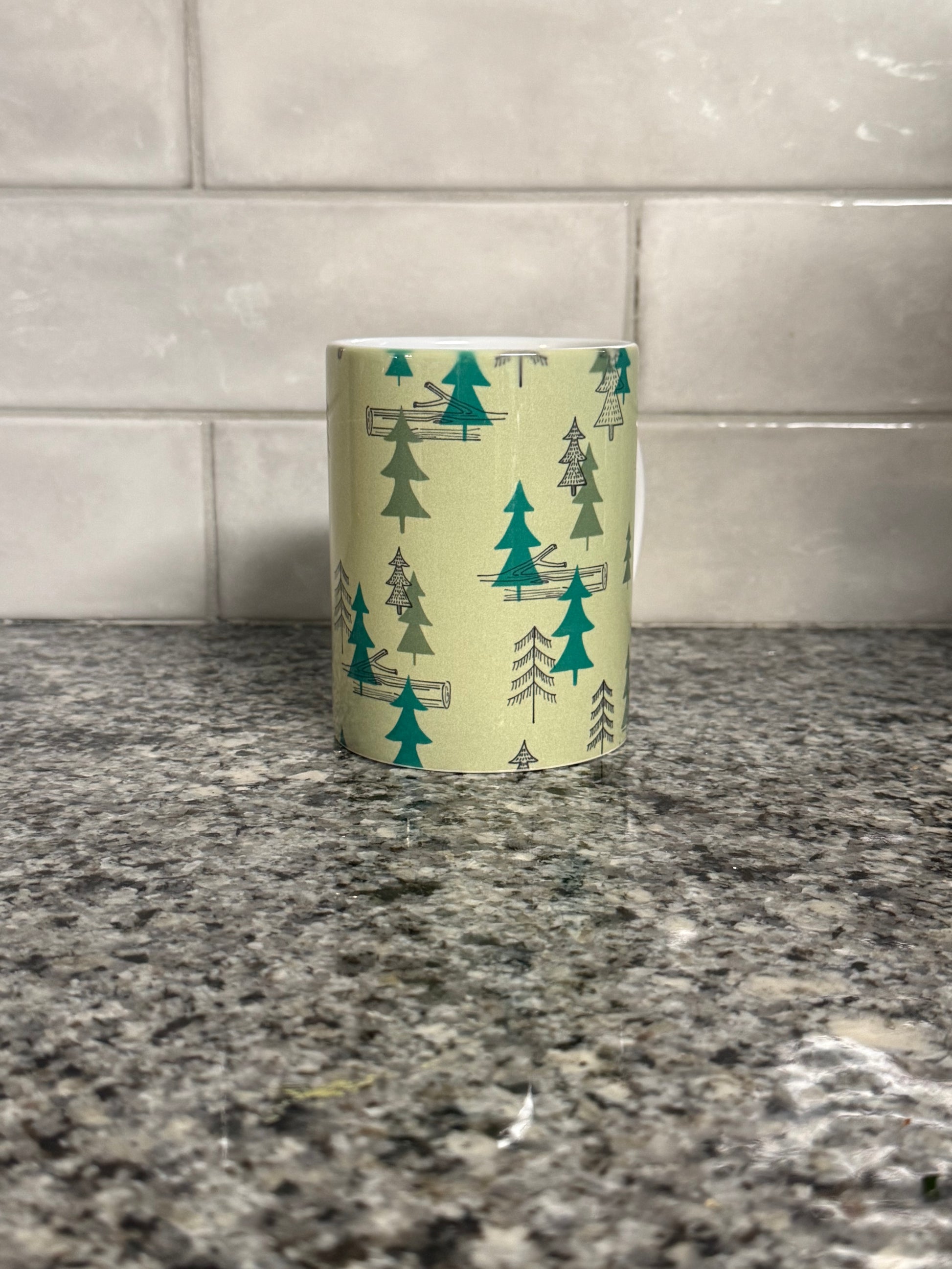 Center view of a 12 oz coffee mug with camping site forest trees design on a granite countertop. Roxzell