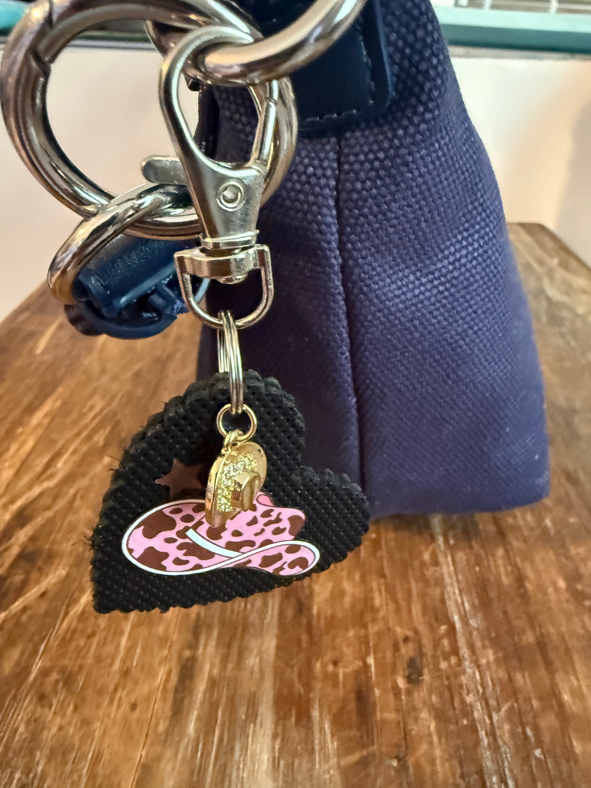 A close-up of a black heart-shaped keychain charm attached to a silver metal key ring and clasp. The bag charm features a pink leopard-print cowboy hat and a small gold cowgirl hat charm, set against a navy fabric bag on a wooden table. Roxzell