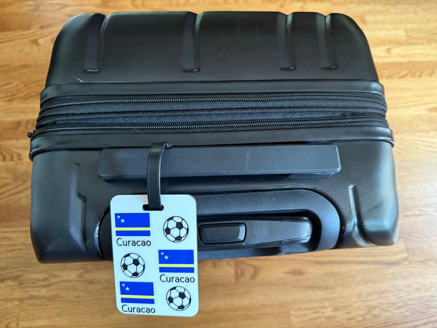 White luggage tag with soccer balls and Curaçao flag featuring blue, yellow stripe, and stars on a black suitcase. Roxzell