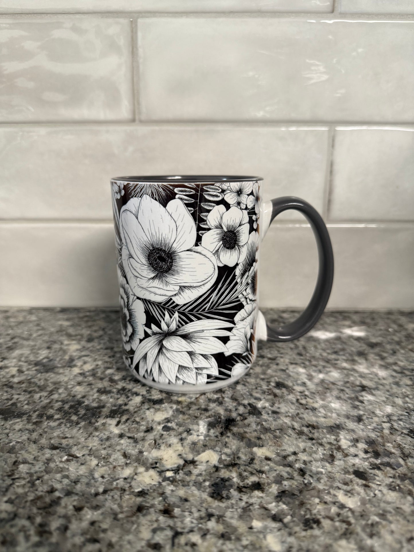 A black ceramic mug with white floral artwork placed on a speckled granite countertop against a tiled wall. Roxzell