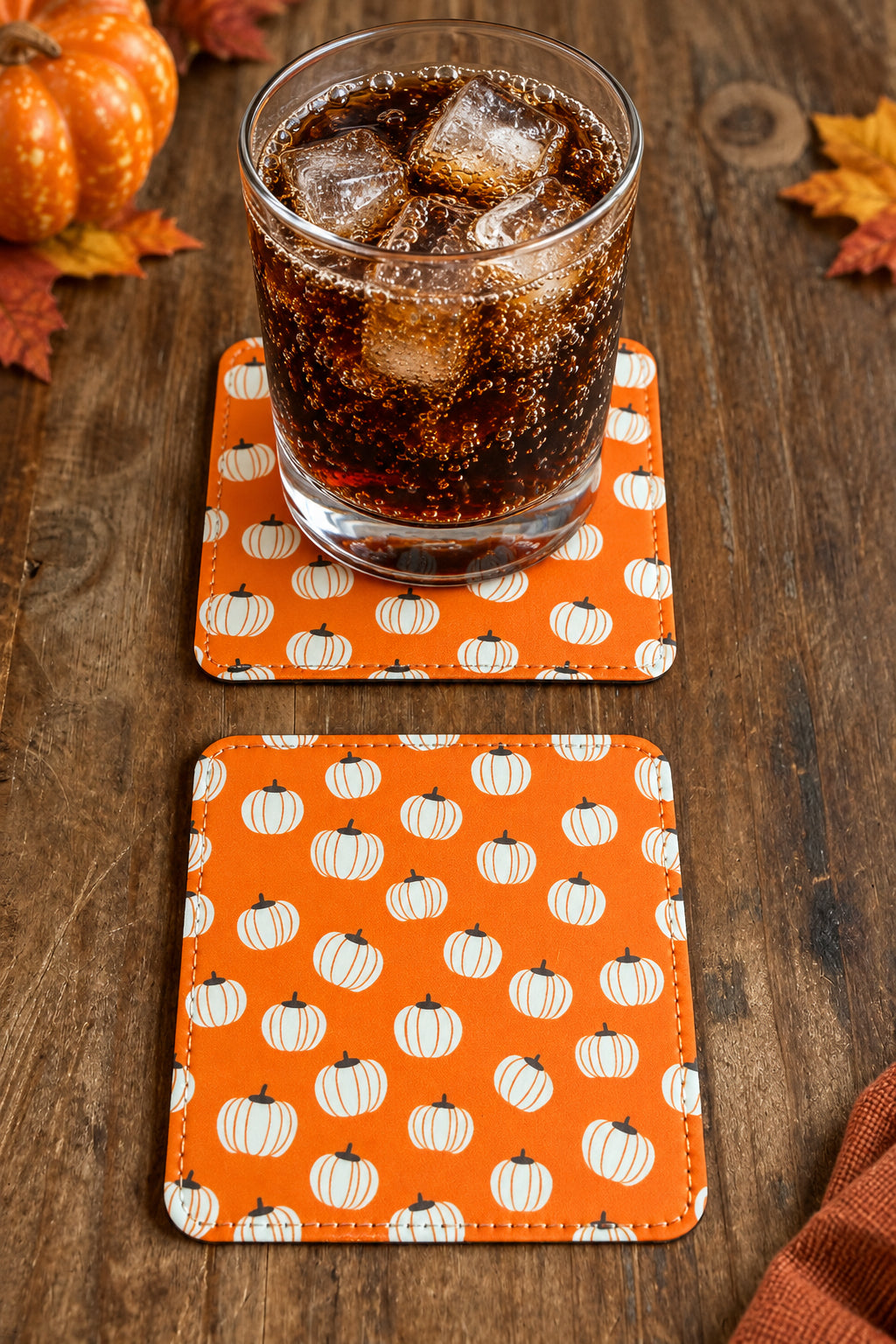 A glass of pop on one of 2 square drink coasters with white pumpkin pattern on a wood table with pumpkin decorations.  Roxzell