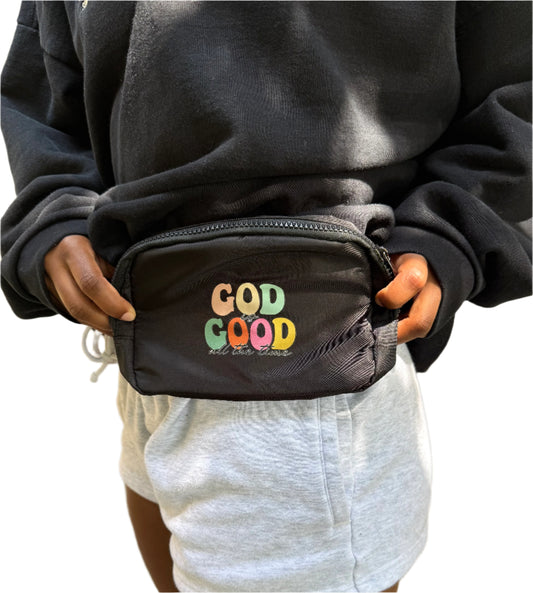 Person holding a black fanny pack/shoulder bag with colorful 'God is Good' text on the front.