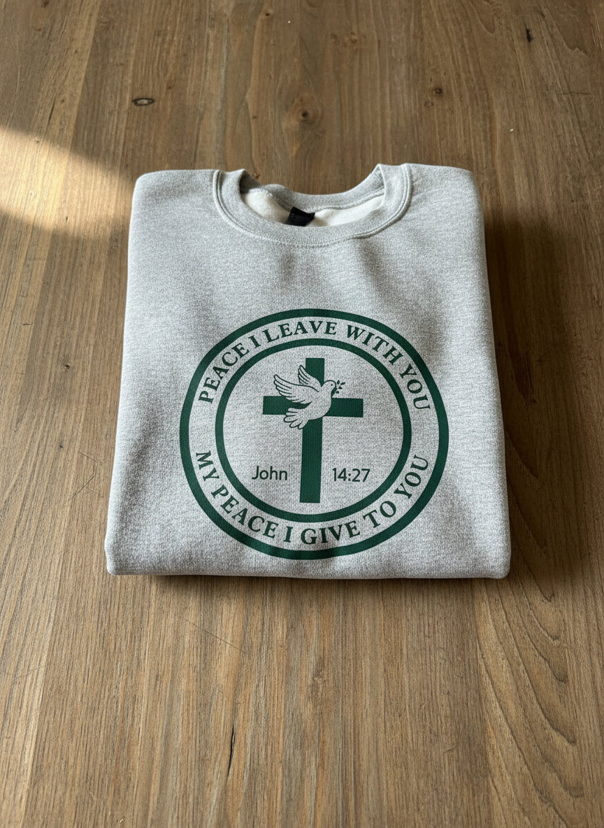 Gray sweatshirt with a green circular design featuring a cross and Peace I Leave With You design on a wood table, Roxzell