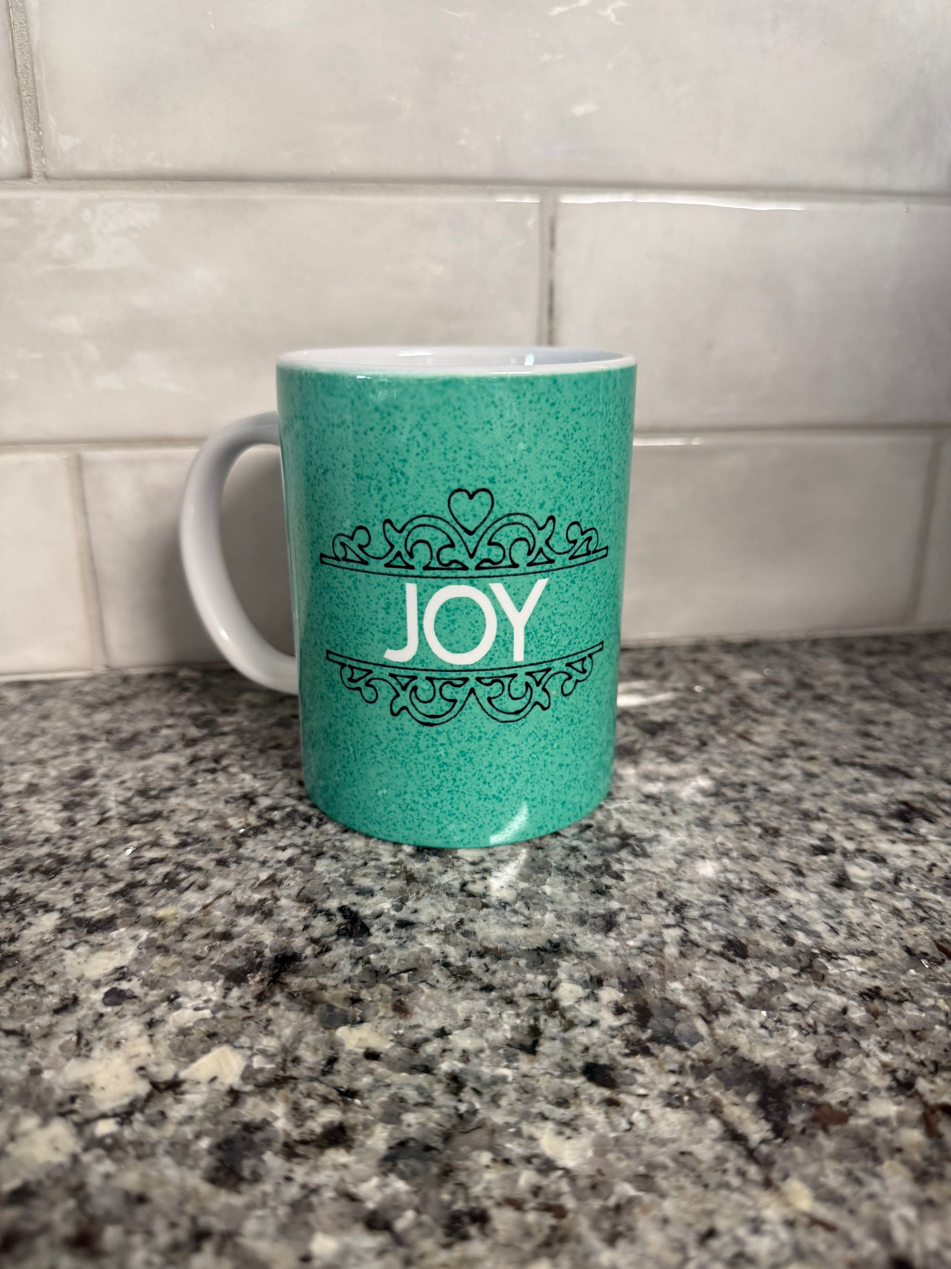 A green ceramic coffee/tea mug with the word 'JOY' written on it, displayed against a kitchen backdrop.