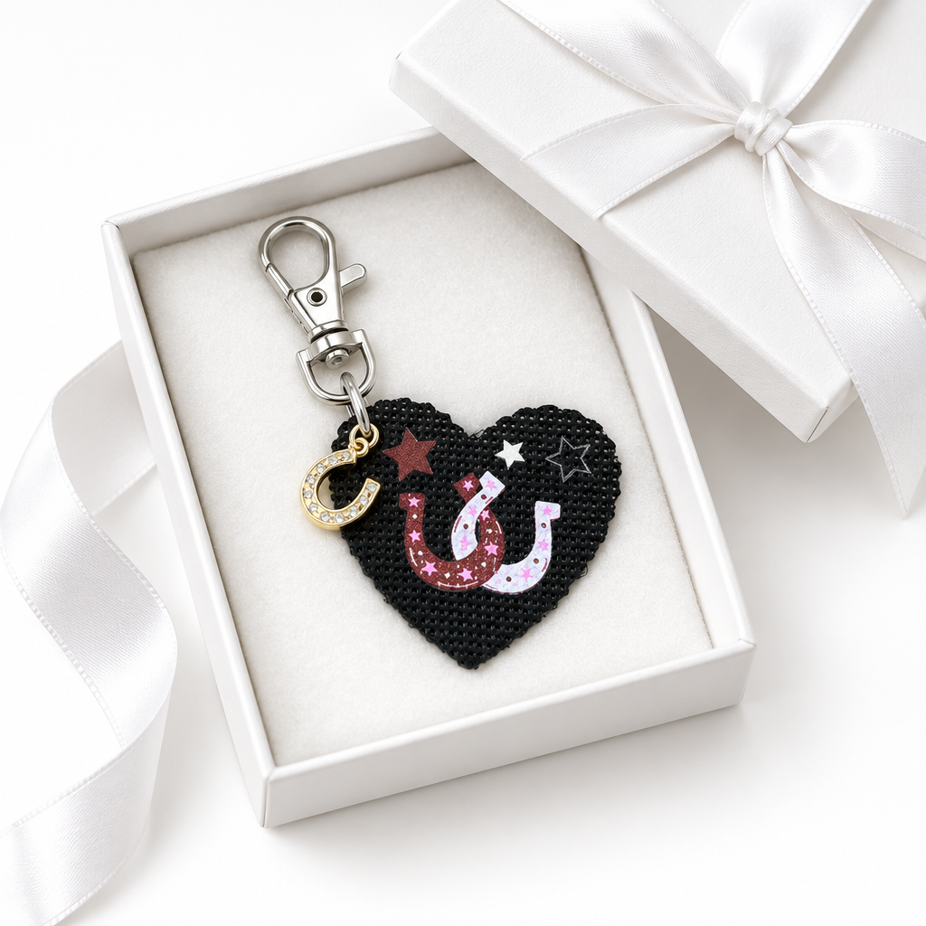 A heart-shaped bag charm with a black background, with brown and pink horseshoes, white stars, and a gold horseshoe charm accent, presented in an open white gift box with a white ribbon bow. Roxzell