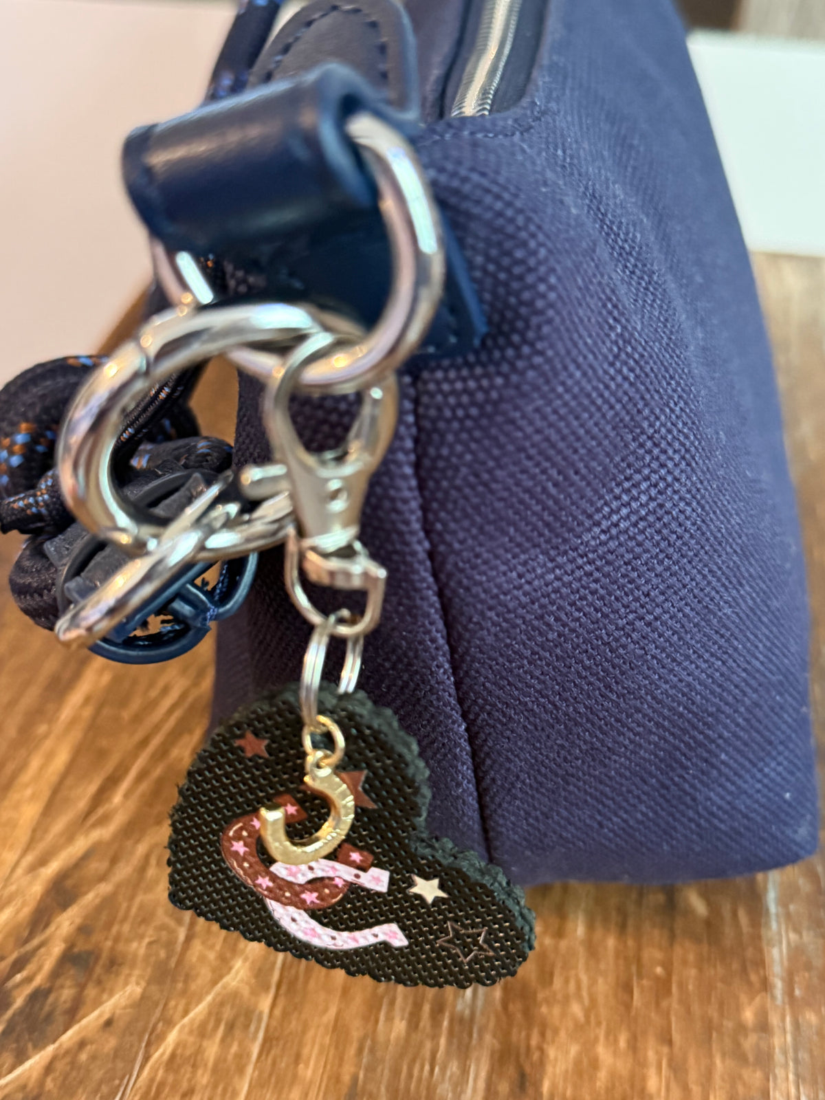 Close-up of a dangling heart-shaped bag charm featuring a pink and gold crescent moon design attached to a navy blue fabric handbag, resting on a wooden surface. Roxzell