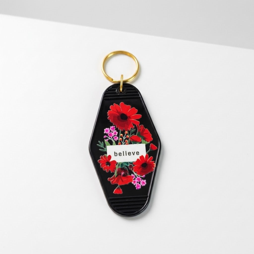 A keychain with a black background, vibrant red and pink flowers, a small white banner reading 'believe', and a gold metal key ring, shown on a clean white surface. Roxzell