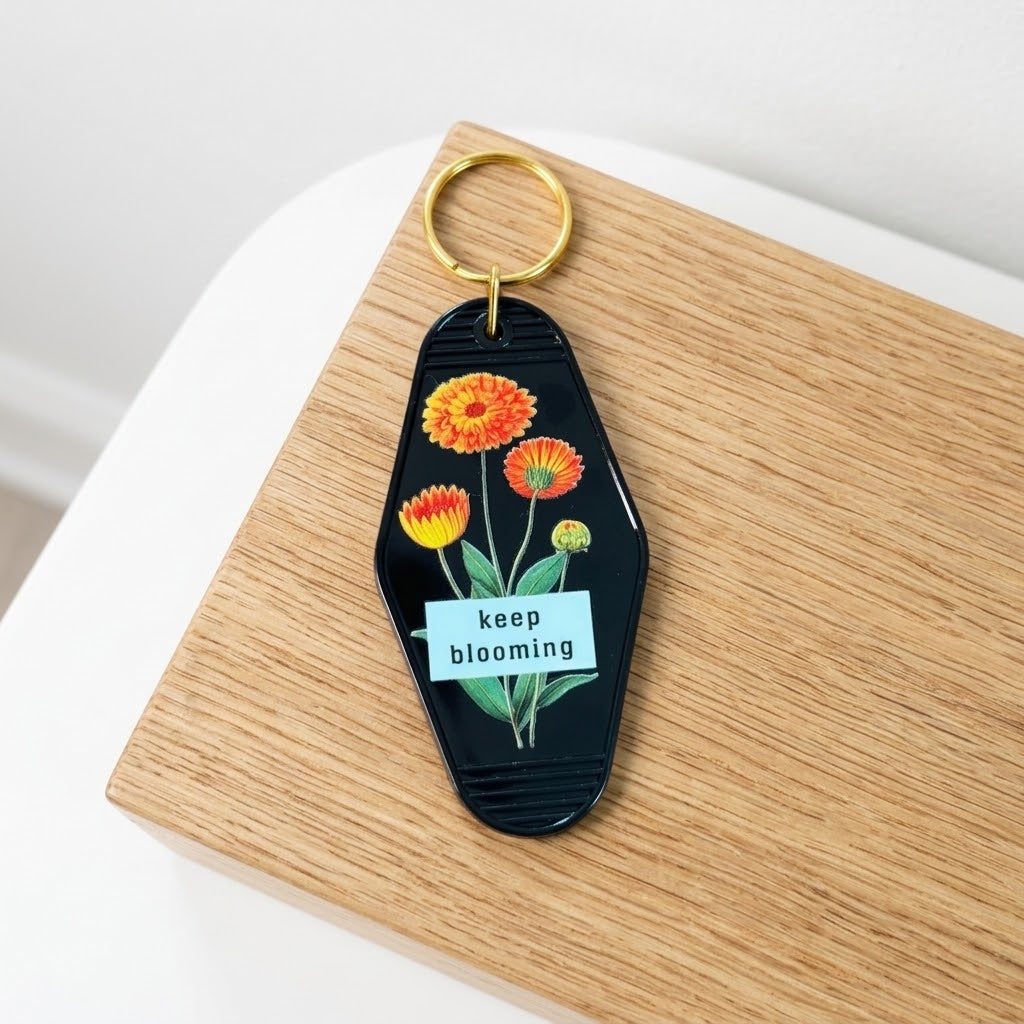 A black acrylic keychain with a gold split ring, featuring an illustration of orange and yellow flowers, green leaves, and a white banner that reads 'keep blooming'. The keychain is resting on a light wood block against a white background. Roxzell