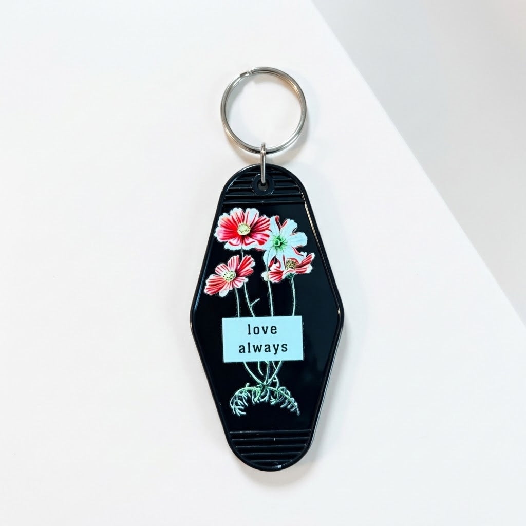 A black acrylic keychain with a silver metal key ring, decorated with red and white flowers, green stems, and a white label that reads 'love always'. Roxzell