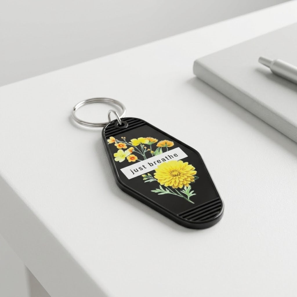 A black keychain with a silver split ring, featuring yellow and orange flowers and the text 'just breathe' on a white label, resting on a white desk next to a gray notebook. Roxzell