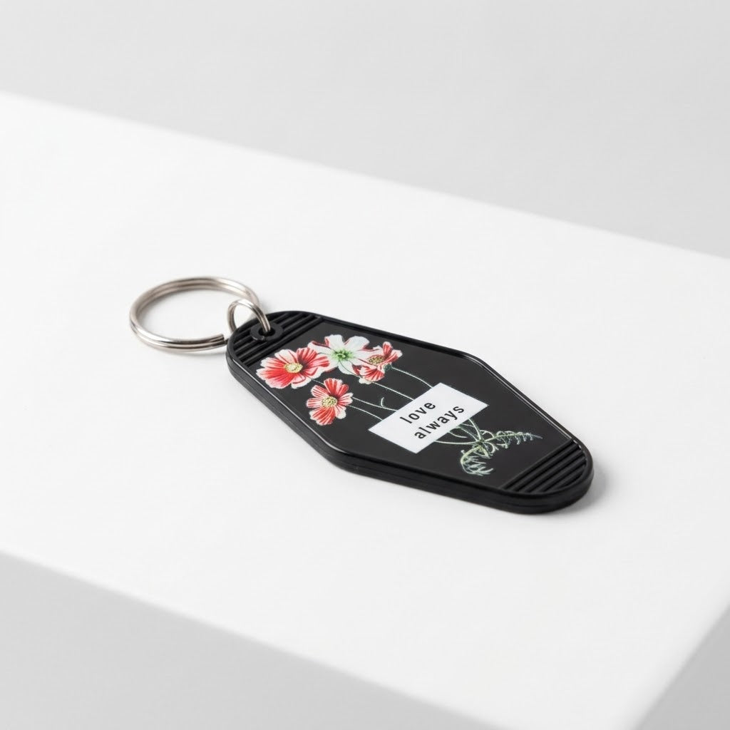 A black keychain with a silver metal split ring, featuring a white label that reads 'love always' in black text, decorated with red and pink flowers and green leaves, resting on a white surface. Roxzell
