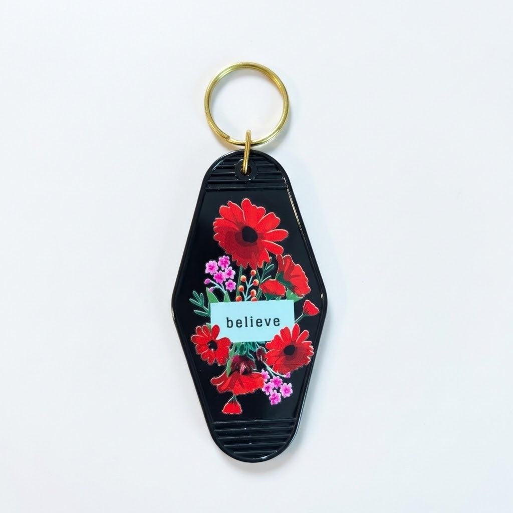 A coffin-shaped keychain with a black ribbed background, decorated with red and pink flowers, green leaves, and a small white banner reading 'believe'. It has a gold metal key ring at the top and is photographed on a clean white background. Roxzell