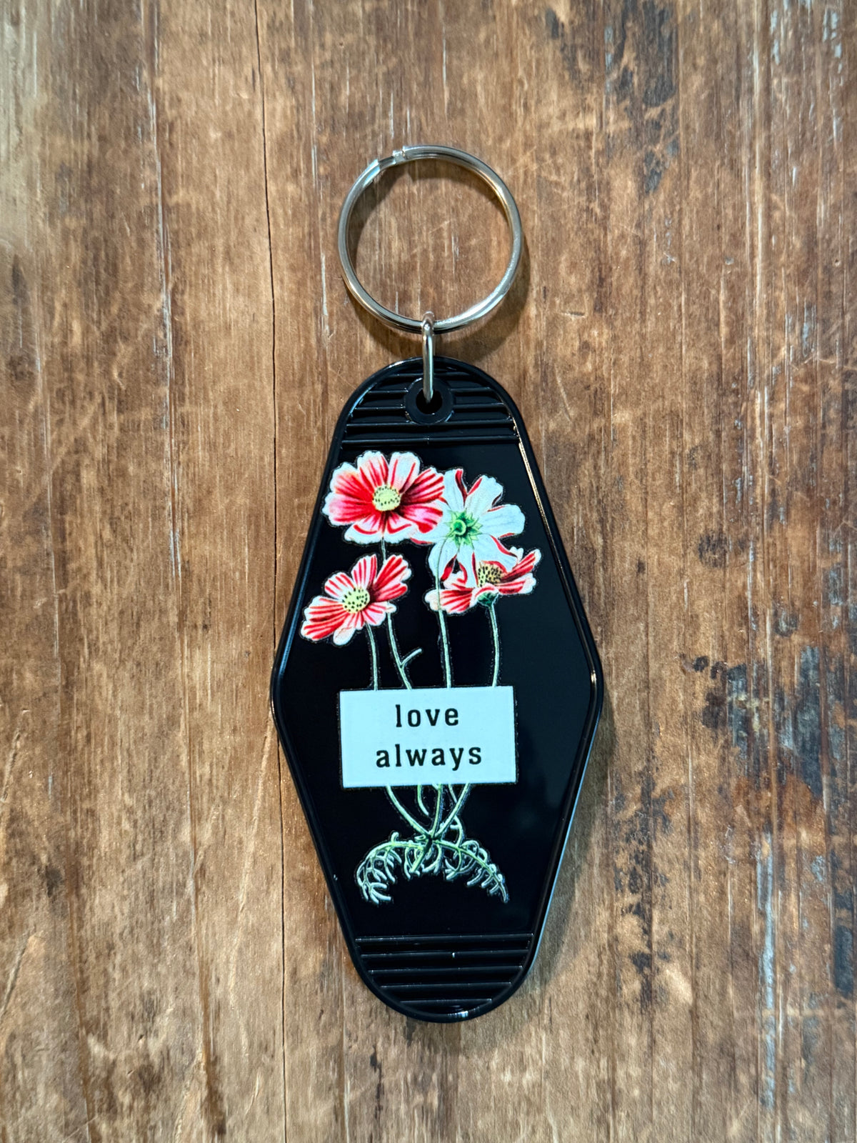 A black keychain with a silver metal split ring, featuring a white label that reads 'love always' in black text, decorated with red and pink flowers and green leaves, resting on a wooden surface. Roxzell