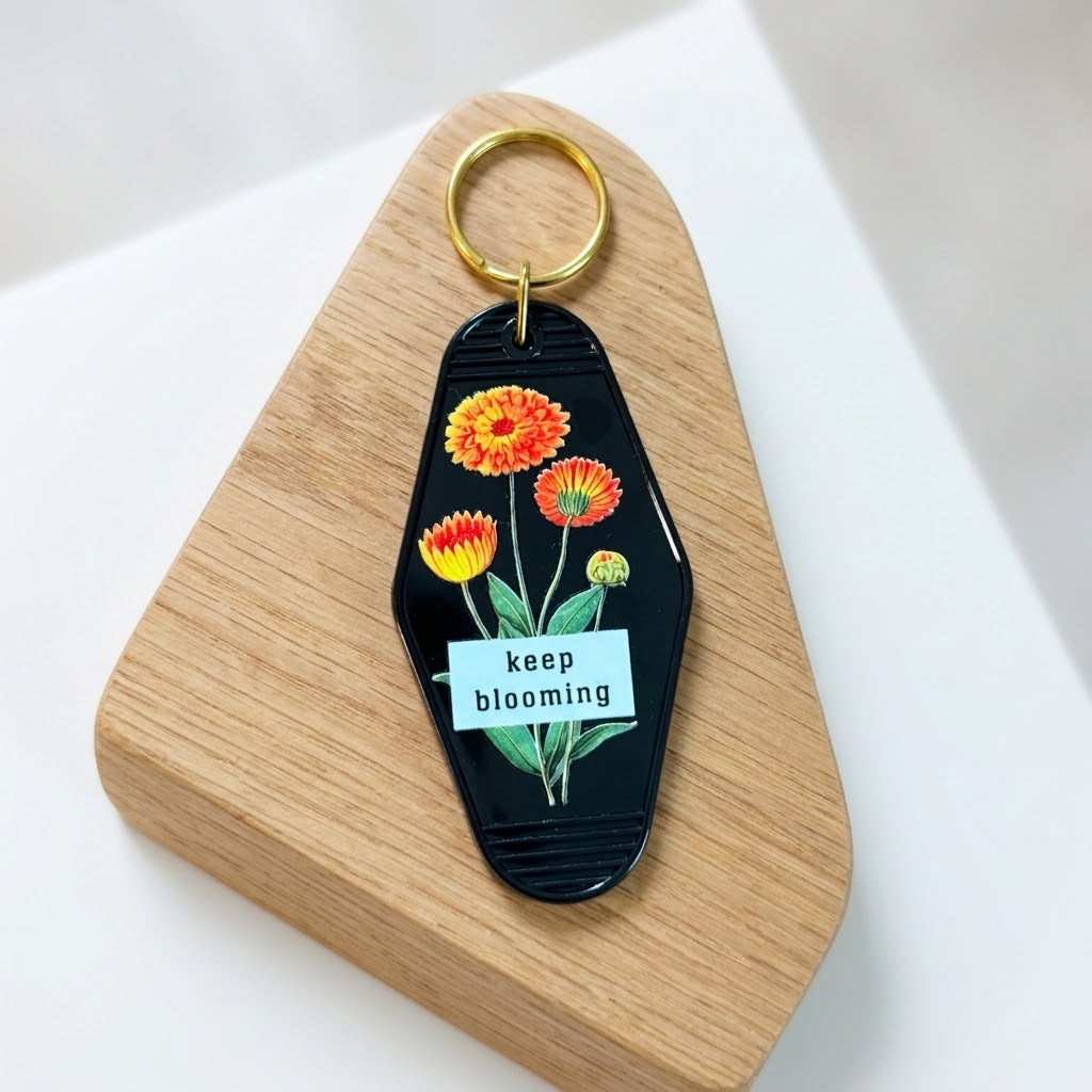 A black acrylic keychain with colorful flower illustrations and a white 'keep blooming' label, attached to a gold key ring and displayed on a wooden stand against a white background. Roxzell