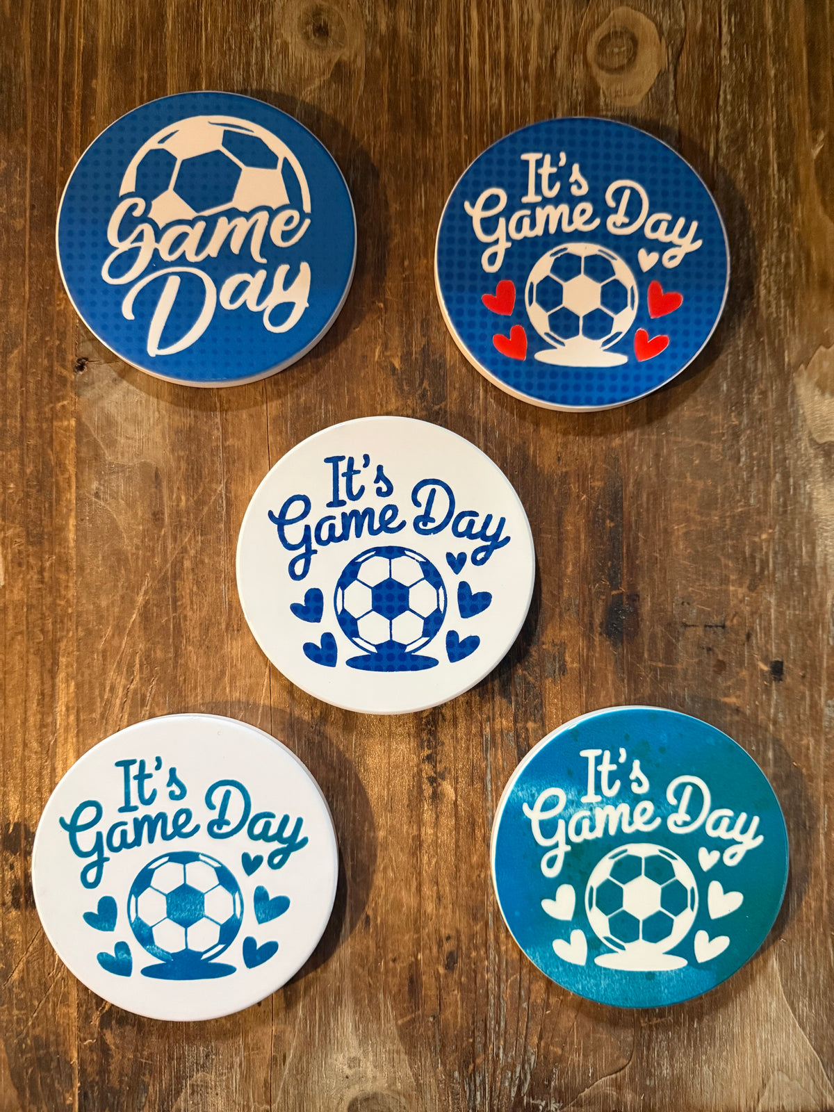 Five round coasters arranged on a rustic wooden surface, each featuring the phrase “It’s Game Day” in cursive white or blue lettering beside a soccer ball graphic surrounded by small hearts. The designs vary in background color—two dark blue, two white, and one teal—with different heart colors (red, blue, or white), creating a cheerful, sporty theme perfect for soccer fans. Roxzell