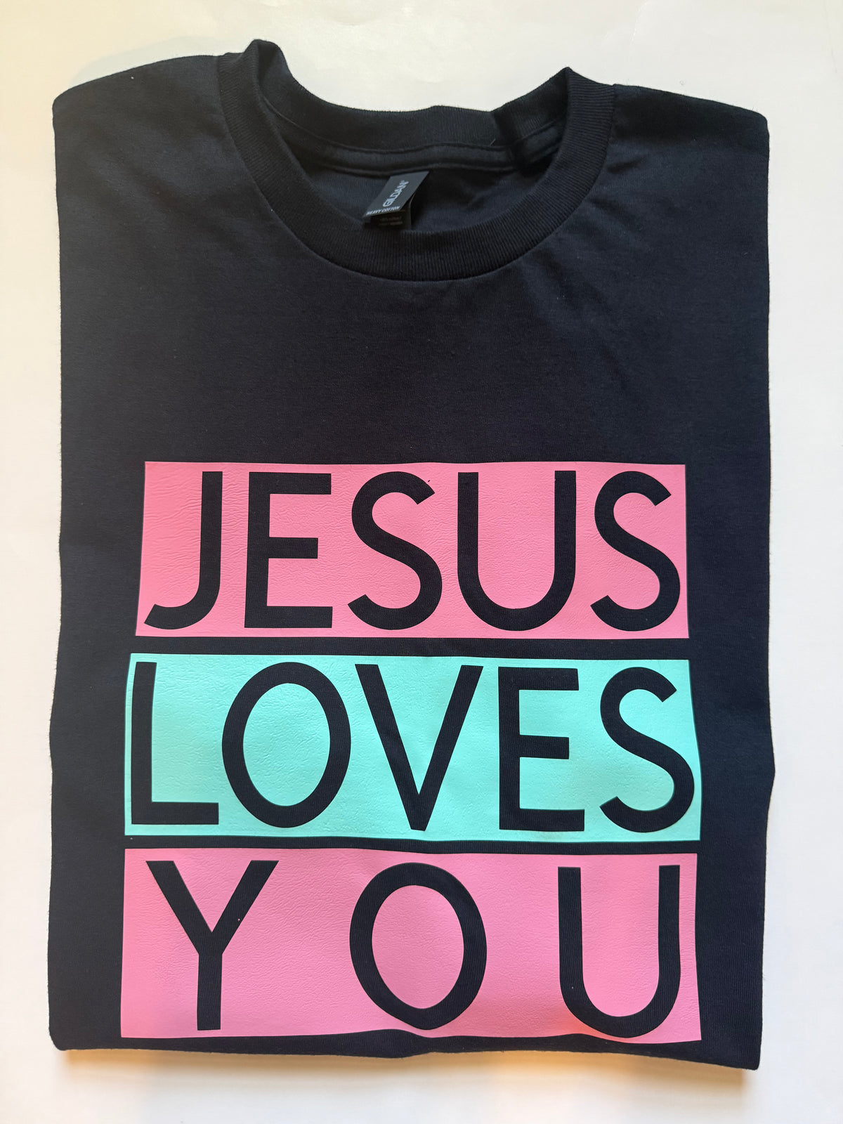 A folded black t-shirt with a graphic print reading 'JESUS LOVES YOU' in bold black letters across three pink and green rectangular blocks, laid flat on a white background. Roxzell