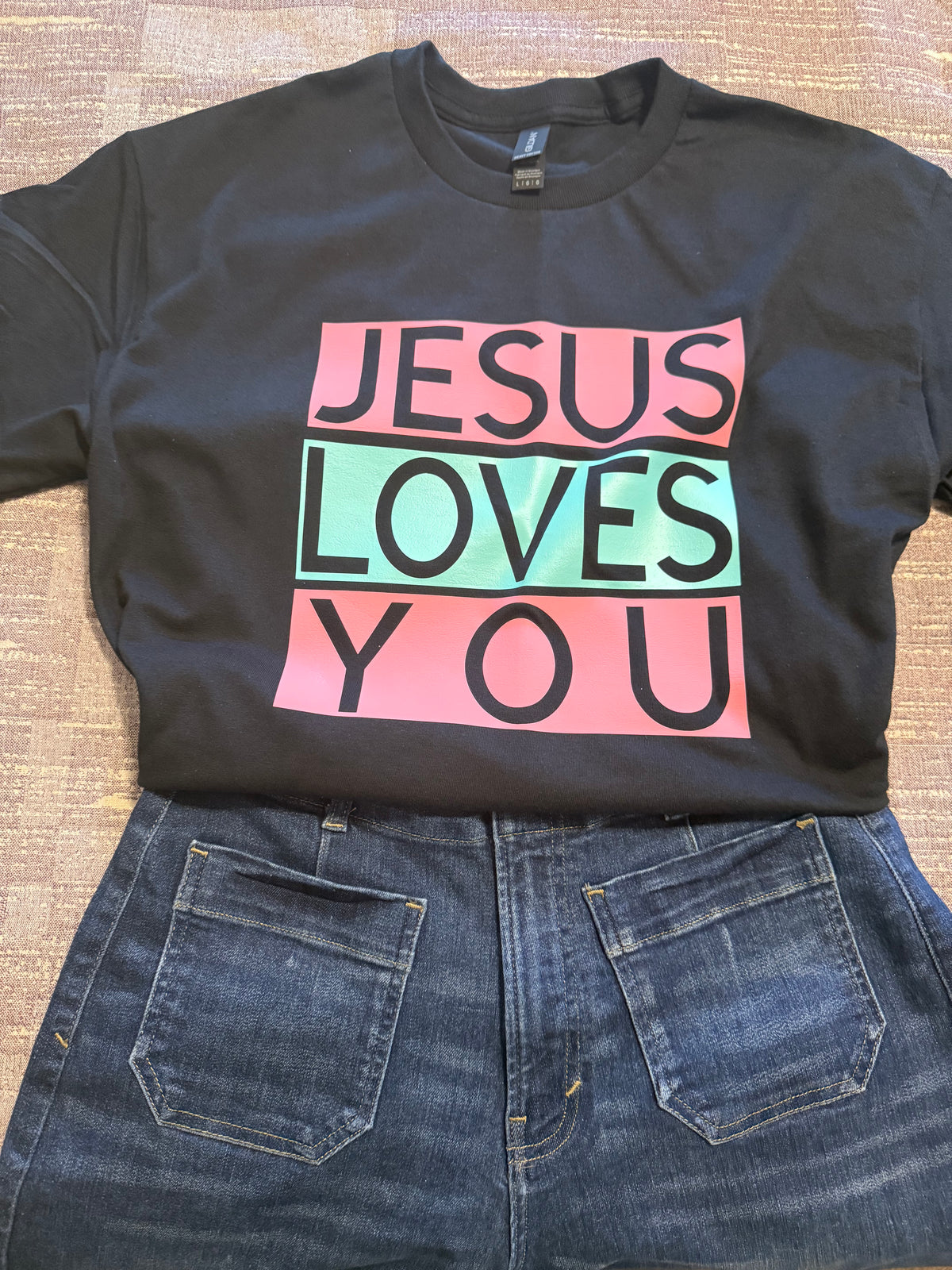 A black short-sleeve graphic T-shirt laid flat on a textured tan surface, paired with blue jeans. The shirt features a large rectangular chest graphic with the words 'JESUS LOVES YOU' in black letters across pink, mint green, and pink blocks. Roxzell