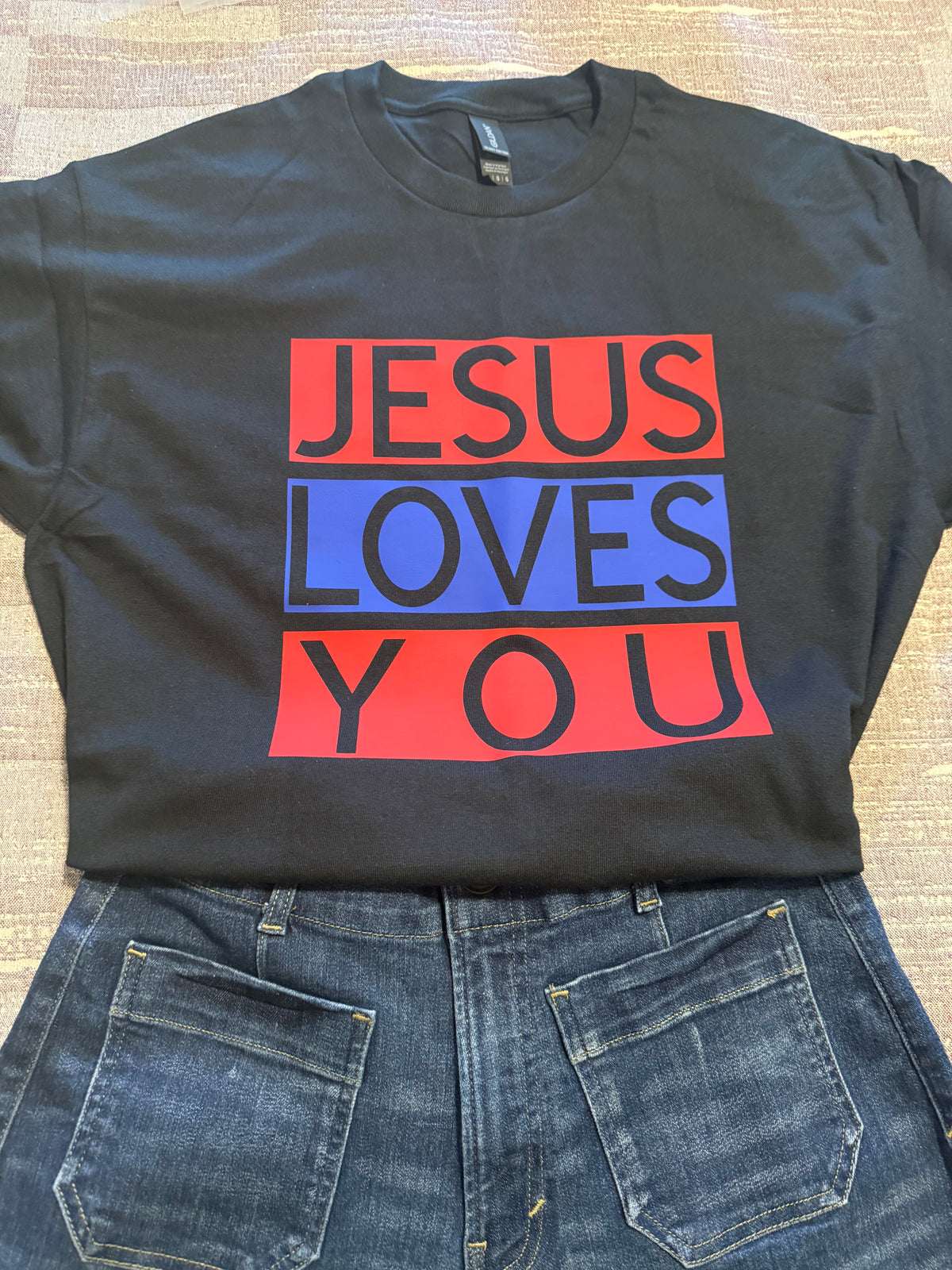 A black graphic t-shirt with the text 'JESUS LOVES YOU' printed in red and blue blocks, styled flat-lay with blue jeans on a textured beige background. Roxzell