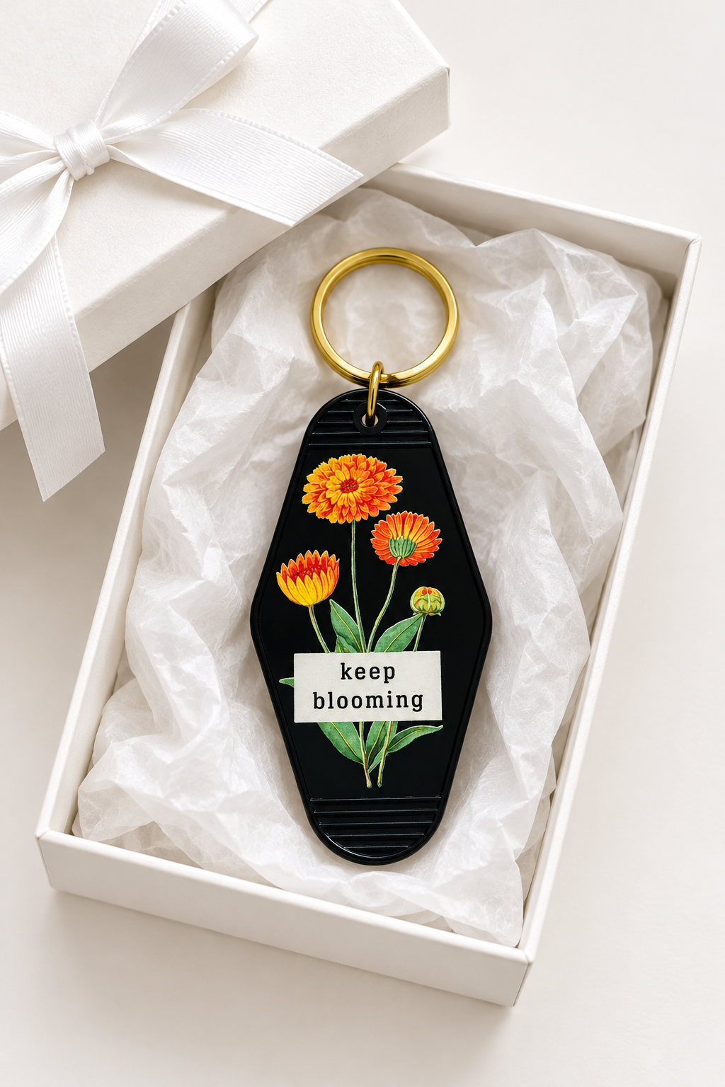 A black acrylic keychain with colorful flowers and the text 'keep blooming' o\in a white gift box with tissue paper and a white ribbon. Roxzell