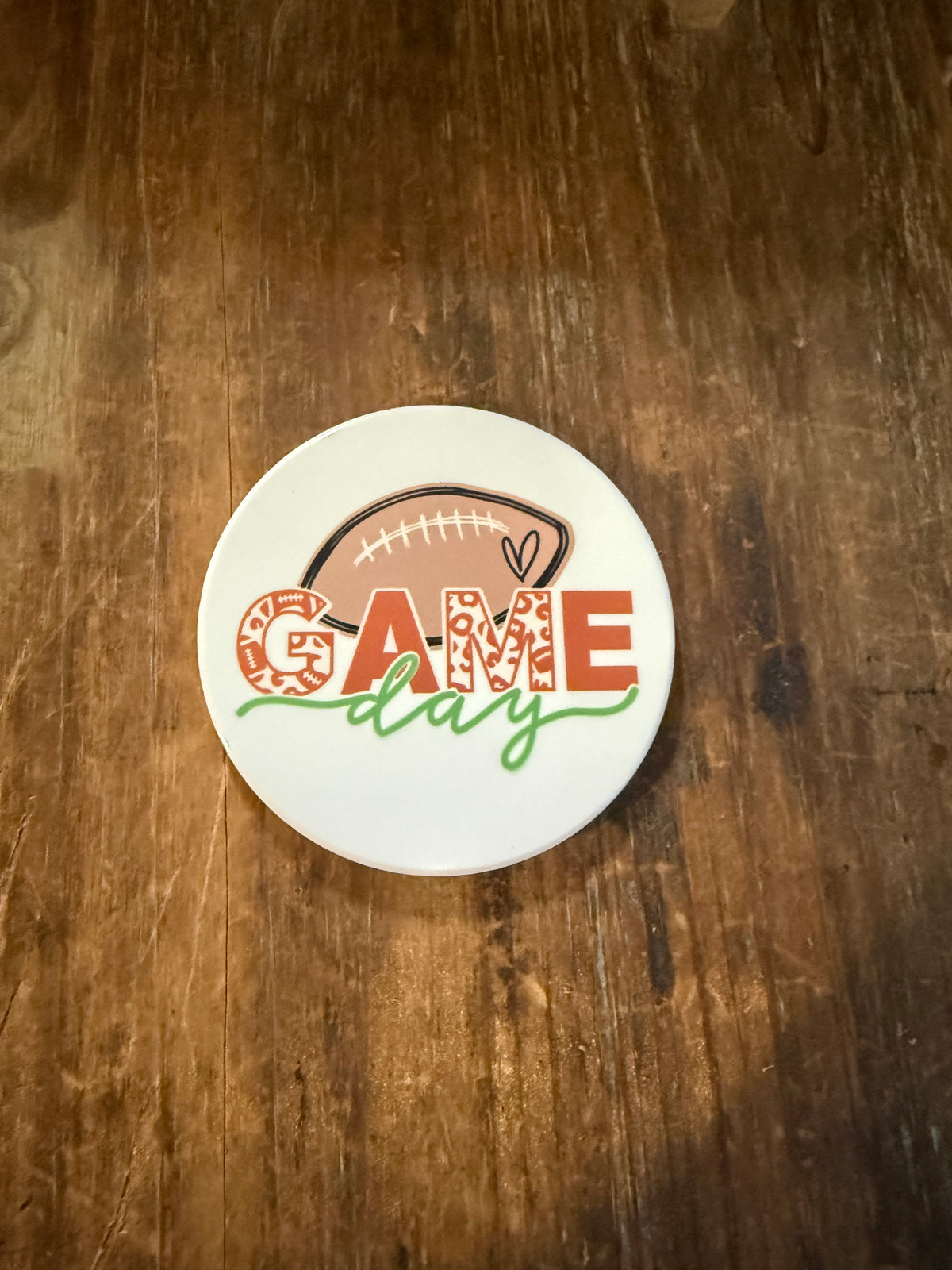 Round white coaster featuring a brown football illustration and the phrase “GAME day.” The word “GAME” is styled in red block letters with a leopard print pattern, while “day” appears in green cursive script. The design sits on a rustic wooden surface, combining sporty and playful aesthetics perfect for football fans. Roxzell
