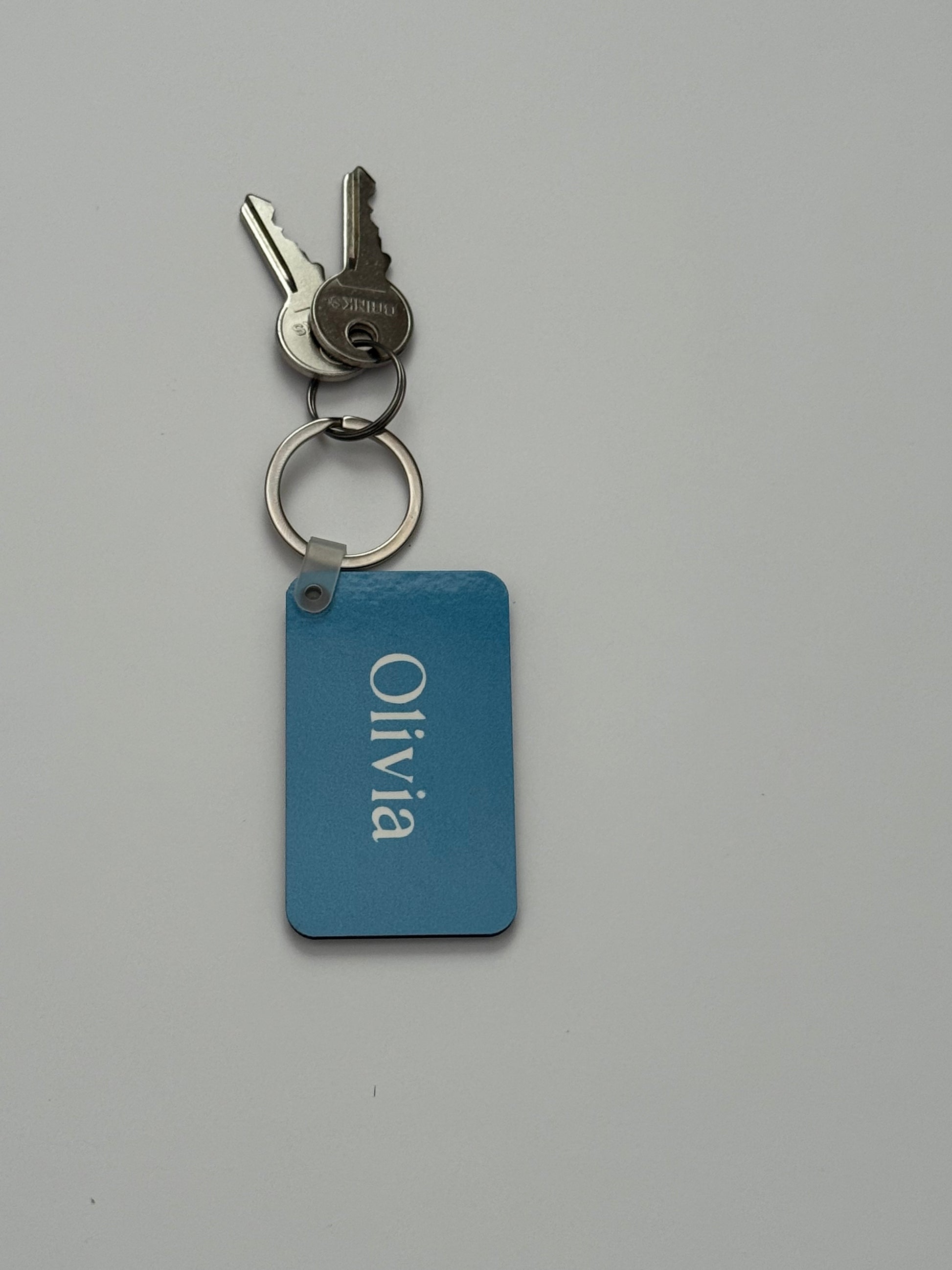 A personalized blue metal key tag with the name 'Olivia' in white script, attached to a split keyring with two keys, on a plain light gray background. Roxzell