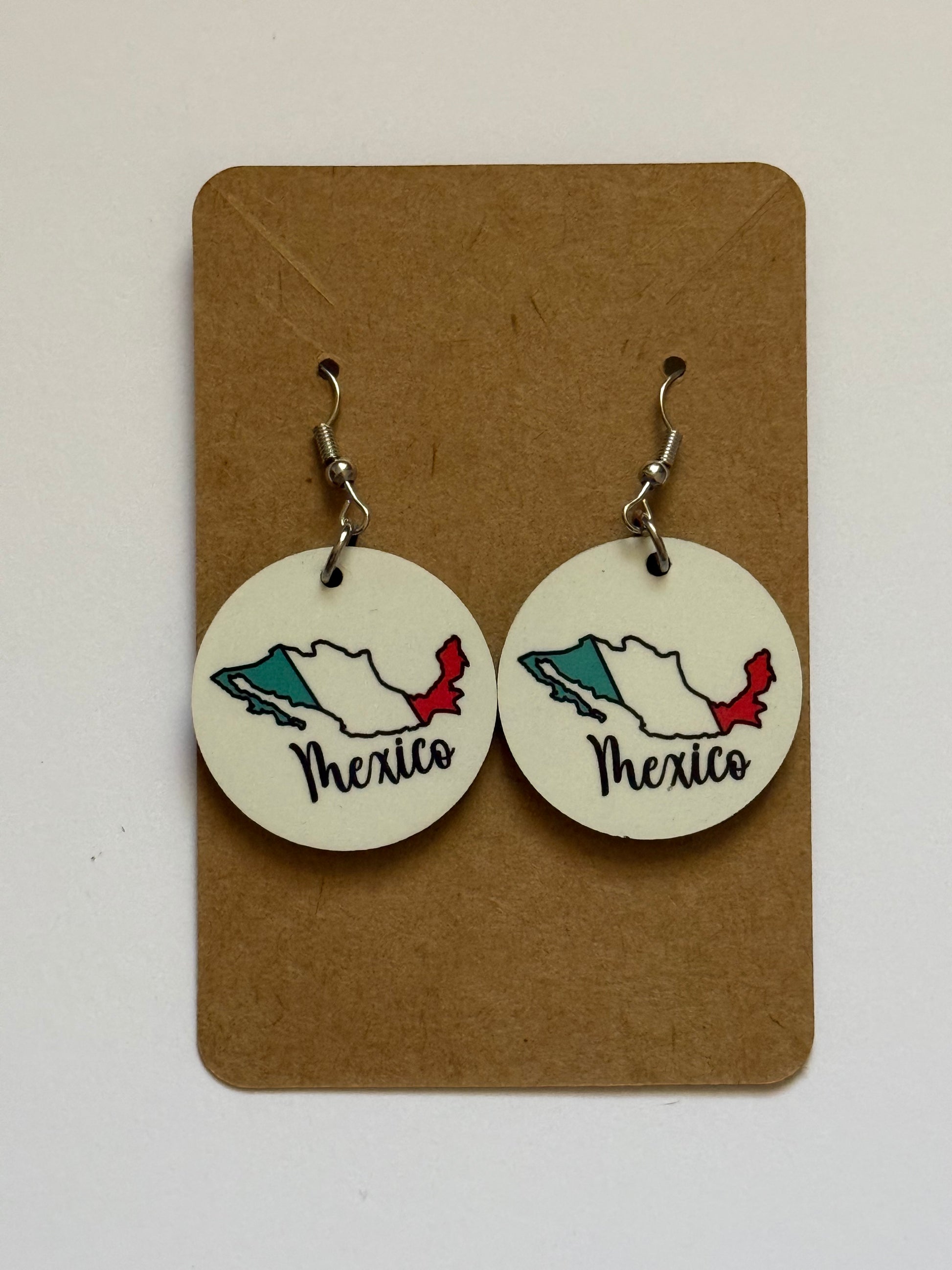 Pair of circular earrings displayed on a brown backing card, each featuring an outline of Mexico colored in green, white, and red to represent the national flag, with the word “Mexico” written in black cursive script beneath the design. Roxzell
