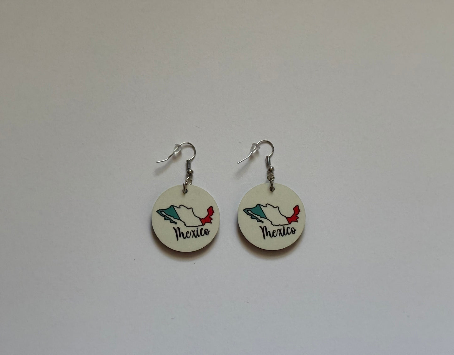 Close‑up of two round earrings attached to a brown card, showing Mexico’s map outline in flag colors — green, white, and red — with “Mexico” written in elegant black cursive lettering on a white background. Roxzell