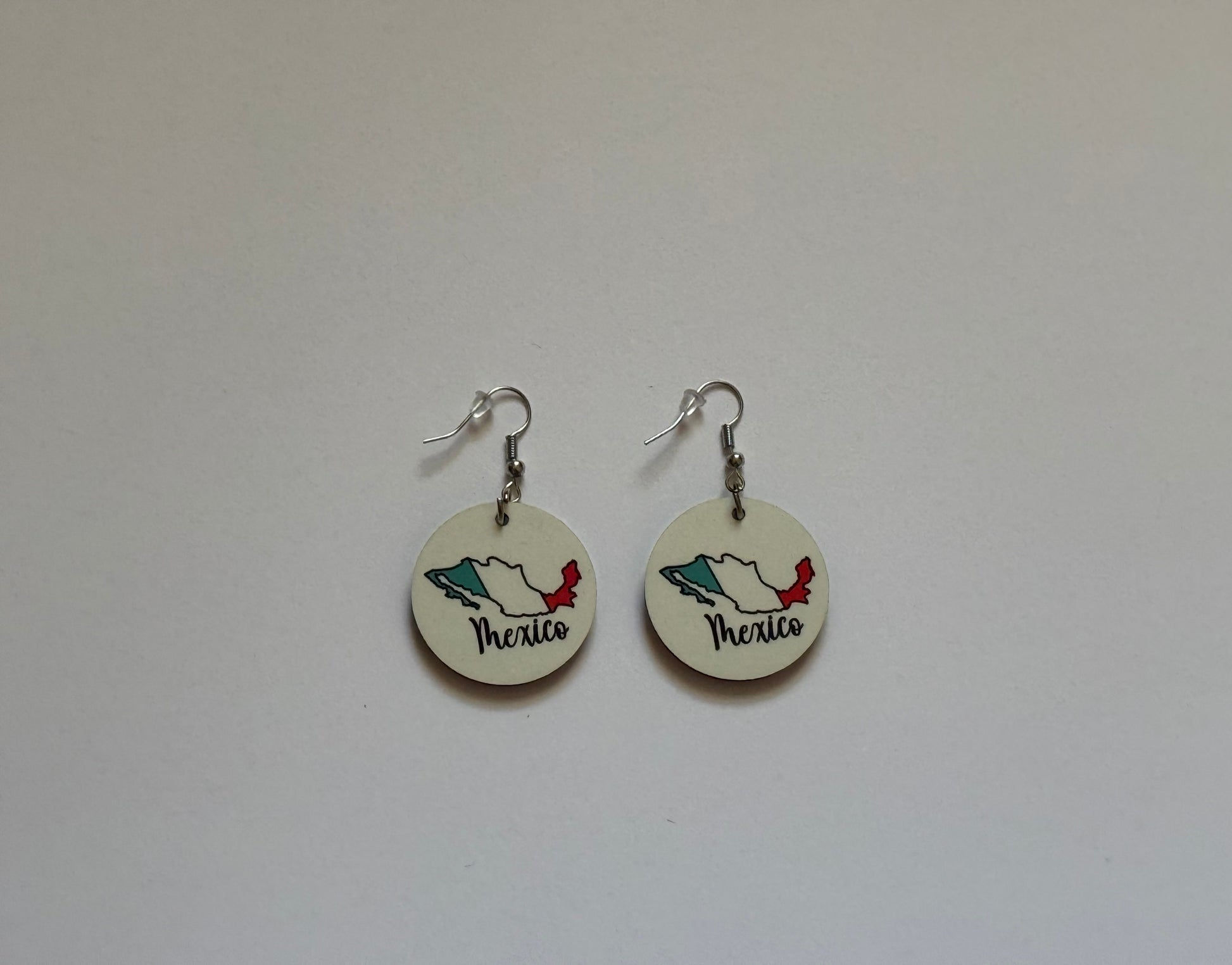 Close‑up of two round earrings attached to a brown card, showing Mexico’s map outline in flag colors — green, white, and red — with “Mexico” written in elegant black cursive lettering on a white background. Roxzell