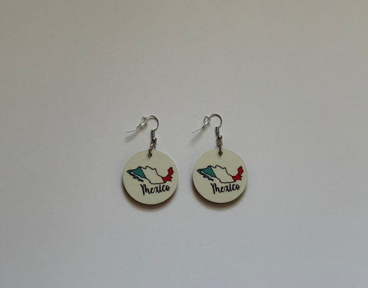 Close‑up of two round earrings attached to a brown card, showing Mexico’s map outline in flag colors — green, white, and red — with “Mexico” written in elegant black cursive lettering on a white background. Roxzell