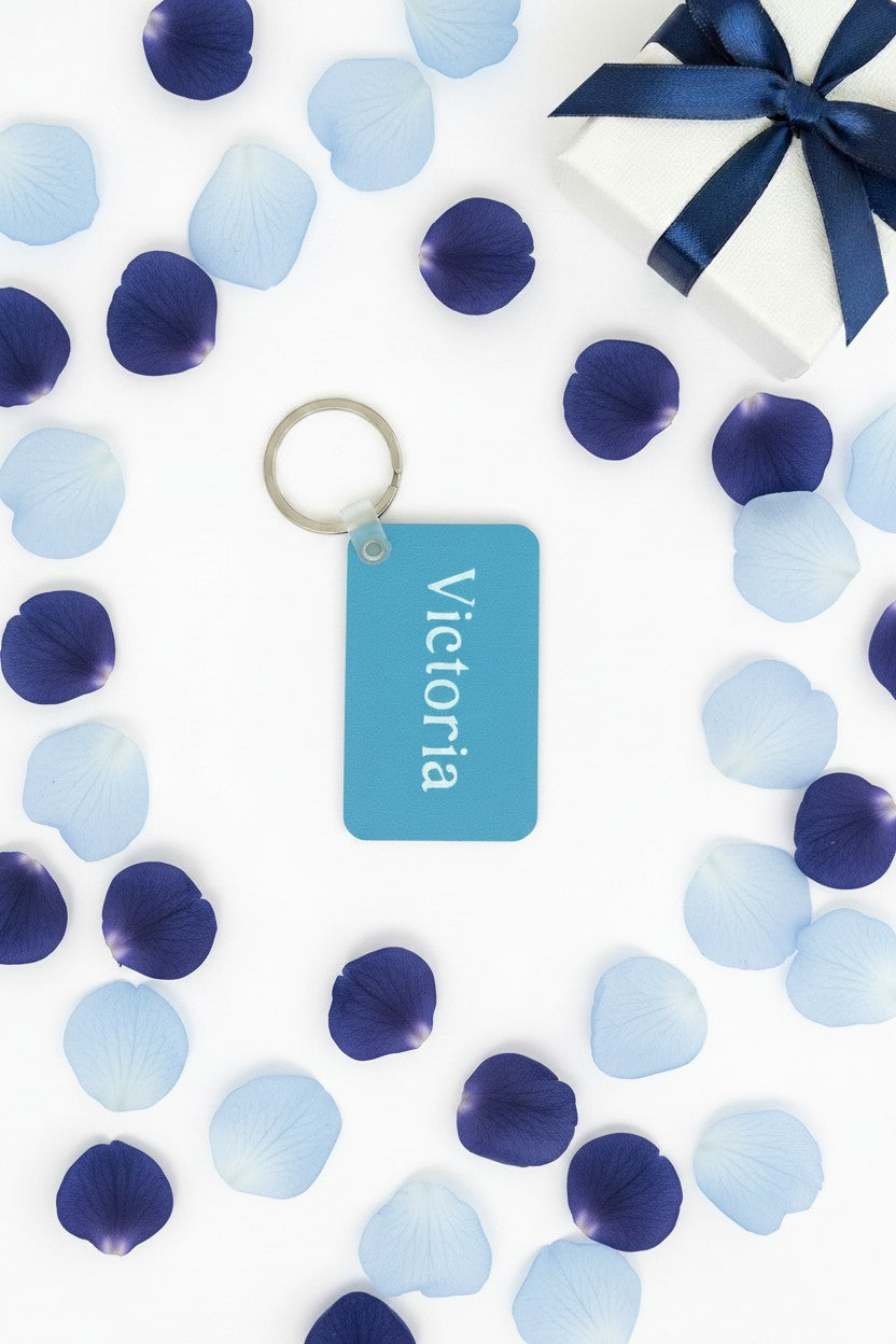 A personalized blue gift tag with the name 'Victoria' printed in white, attached to a silver key ring. The tag is displayed on a white background surrounded by scattered blue and navy petal confetti, with a white gift box and navy ribbon in the top right corner. Roxzell