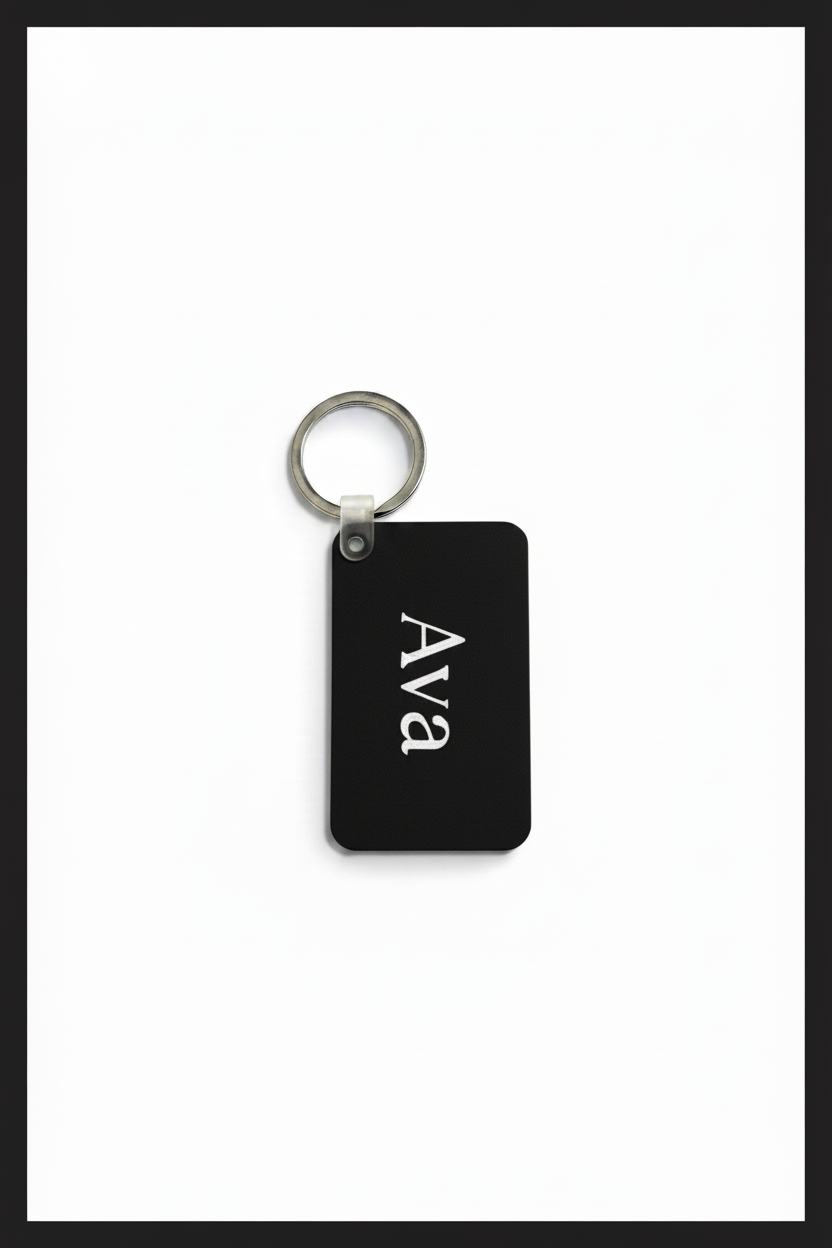 A rectangular black keychain tag with rounded corners, featuring the name 'Ava' printed vertically in white, attached to a silver metal key ring, set against a white background with a black border. Roxzell