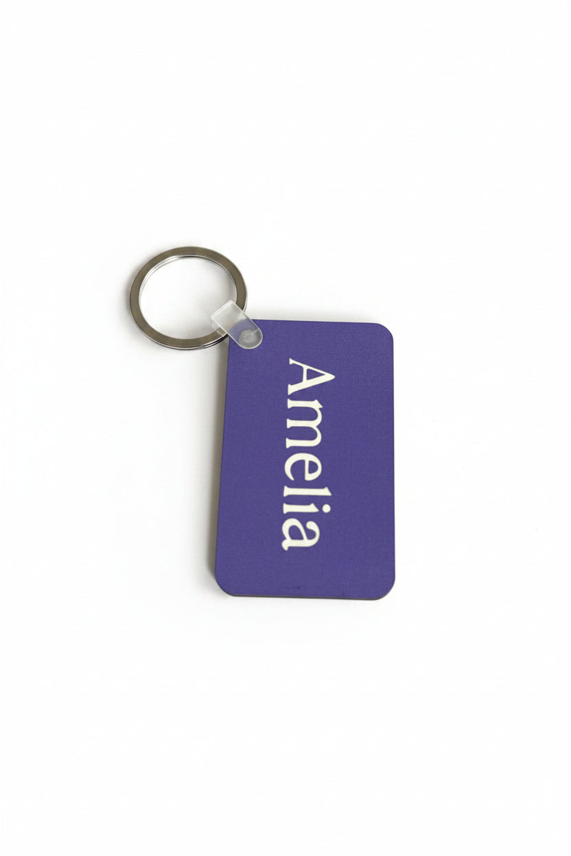 A purple rectangular keychain tag with rounded corners, featuring the name 'Amelia' printed vertically in white. The tag is attached to a silver metal key ring with a clear plastic connector, shown on a plain white background. Roxzell