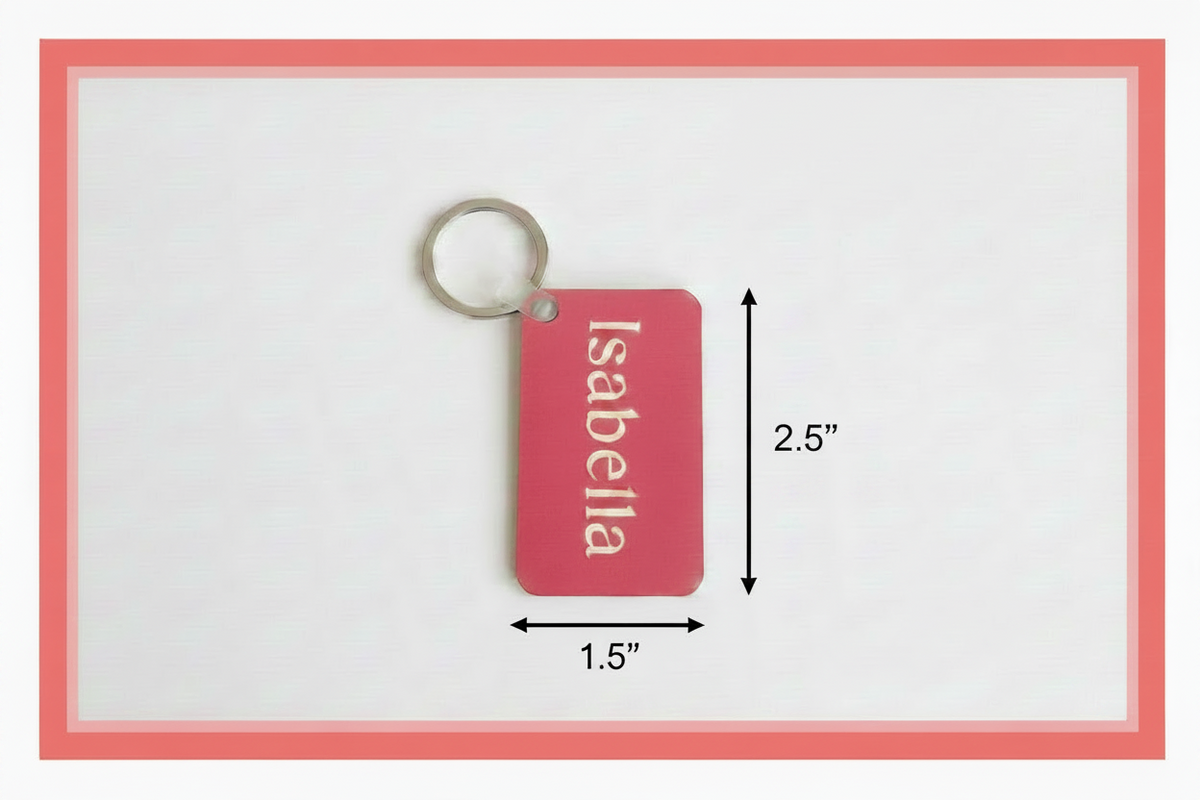 A rectangular pink keychain tag with rounded corners, attached to a silver split ring. The tag has the name 'Isabella' printed vertically in white. A 2.5" dimension arrow is on the right and a 1.5" dimension arrow is on the bottom of the keychain.  Roxzell