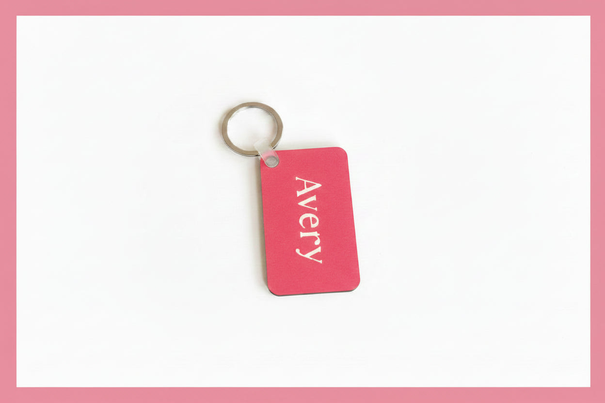 A pink rectangular key tag with rounded corners featuring the white vertical text 'Avery' and attached to a silver metal split ring, shown on a plain white background with pink border. Roxzell