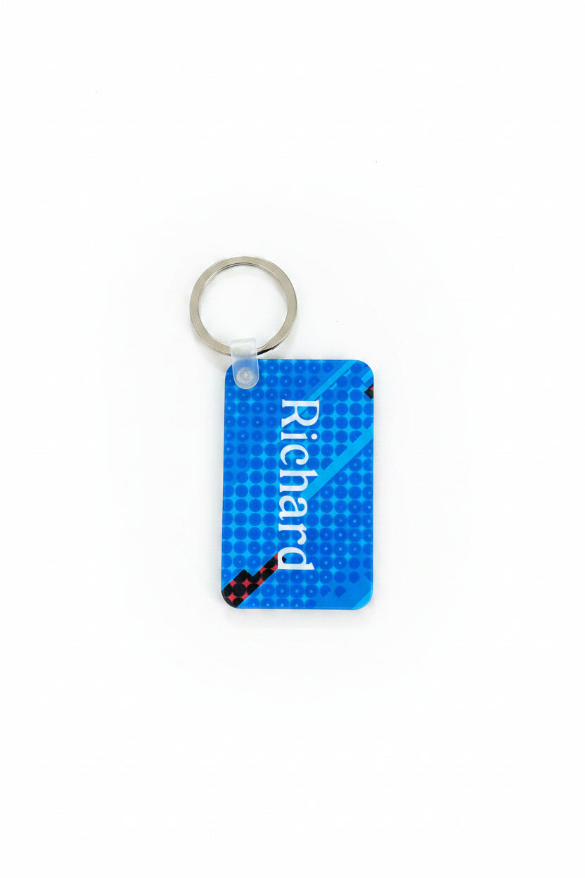 Stylish blue keychain with white “Richard” text and red‑black diagonal accent, displayed on a warm wooden surface.