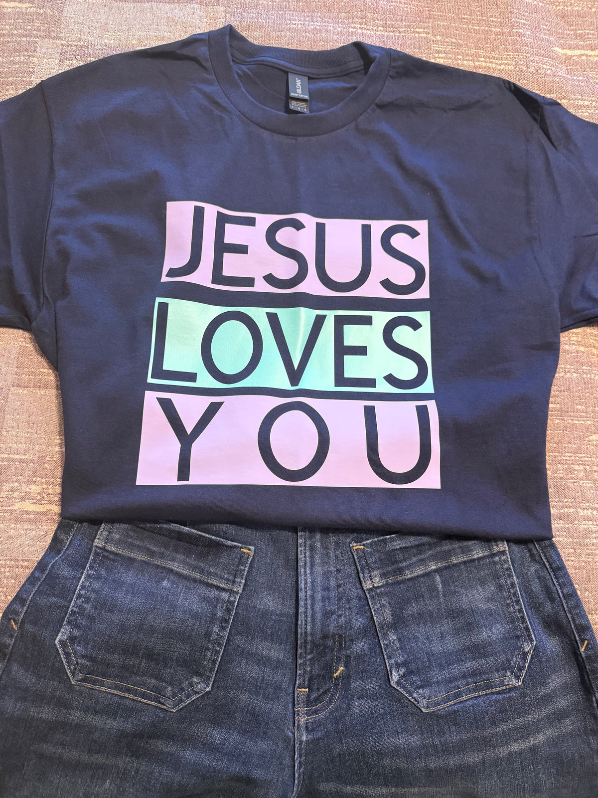 A navy blue graphic t-shirt laid flat on a textured tan surface, paired with blue jeans. The t-shirt features a graphic that reads 'JESUS LOVES YOU' in black block letters across three pastel-colored horizontal bars: light pink, mint green, and light pink. Roxzell
