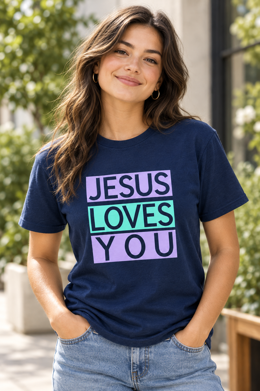 A woman wearing a navy blue graphic t-shirt with the text 'JESUS LOVES YOU' in purple and mint green blocks, paired with light wash blue jeans, standing outdoors in front of blurred green foliage. Roxzell
