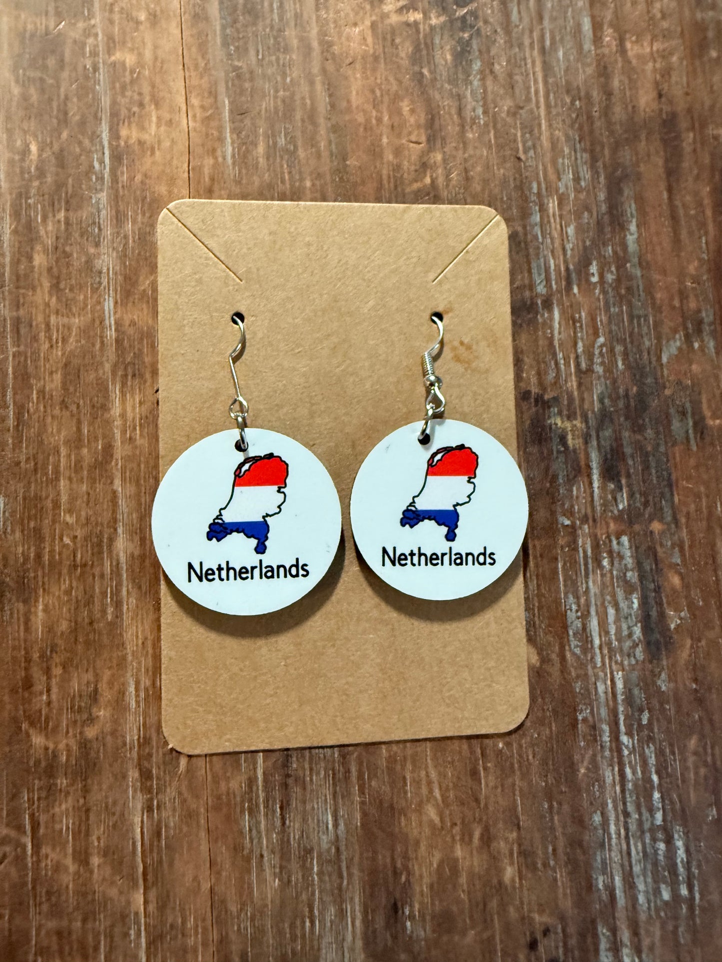 A pair of Netherlands-themed earrings with red, white, and blue map design, presented on a brown cardboard backing over a wooden surface. Roxzell