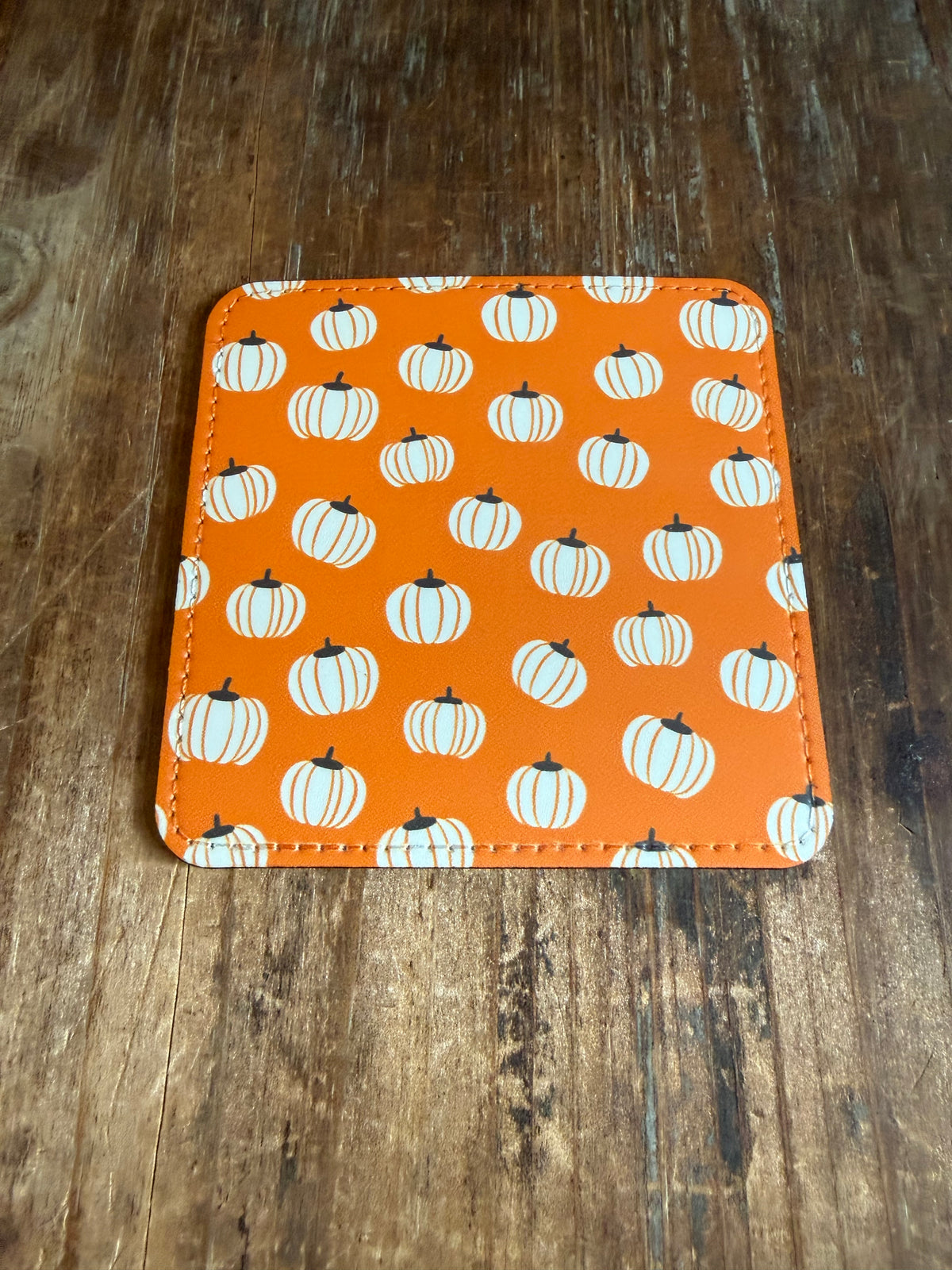 An orange square coaster with rounded corners, featuring a repeating pattern of white pumpkins with black stems, placed on a dark wood surface. Roxzell
