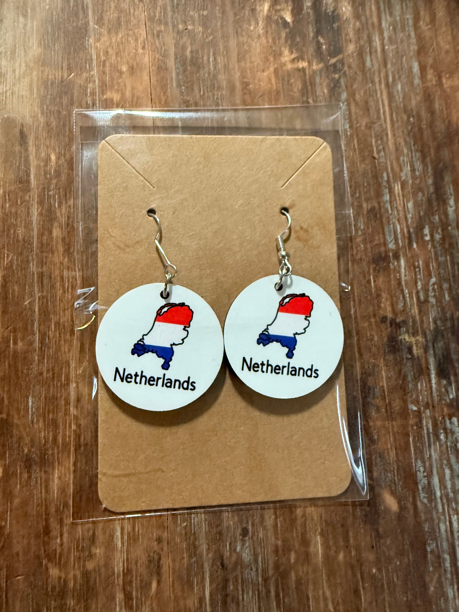 Netherlands map earrings in red, white, and blue, attached to a cardboard backing and sealed in clear plastic packaging on a wooden table.