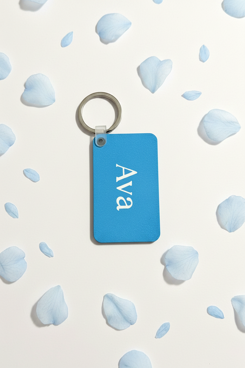 A rectangular blue keychain personalized with the name 'Ava' printed in white script, attached to a silver metal key ring, displayed on a white background scattered with light blue flower petals. Roxzell