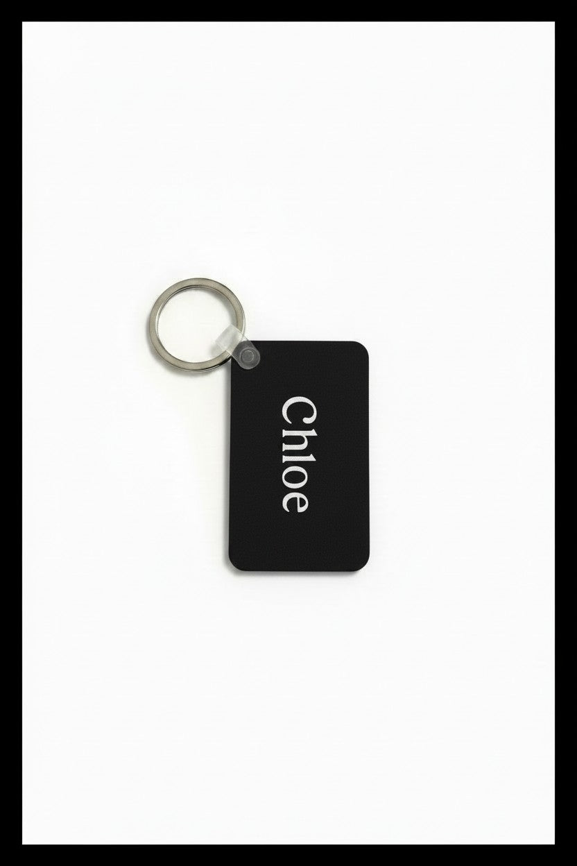 A black rectangular key tag with rounded corners and a silver metal key ring, featuring the name 'Chloé' printed vertically in white on the front, set against a plain white background. Roxzell