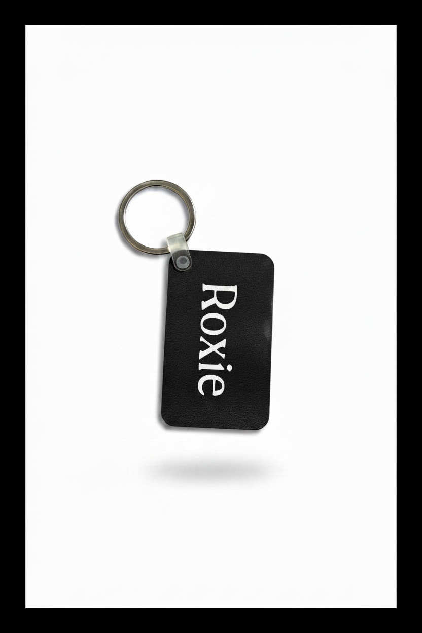 A black rectangular keychain with the name 'Roxie' printed vertically in white serif letters, attached to a silver metal split ring, shown on a white background with a soft drop shadow.