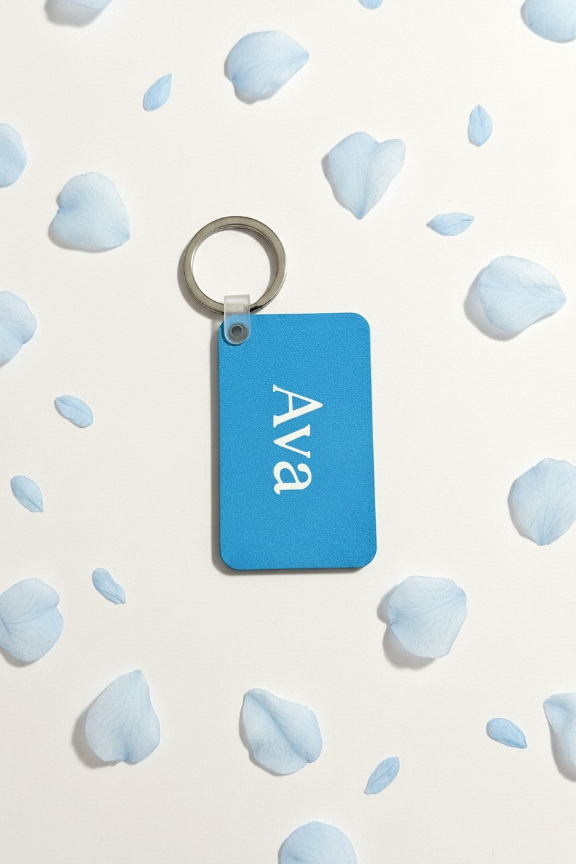 A rectangular blue keychain with the name 'Ava' printed in white script, attached to a silver metal key ring, displayed on a white background scattered with light blue flower petals. Roxzell