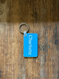 A blue rectangular keychain tag with the name 'Charlotte' printed vertically in white, attached to a silver metal key ring, resting on a dark wood surface. Roxzell
