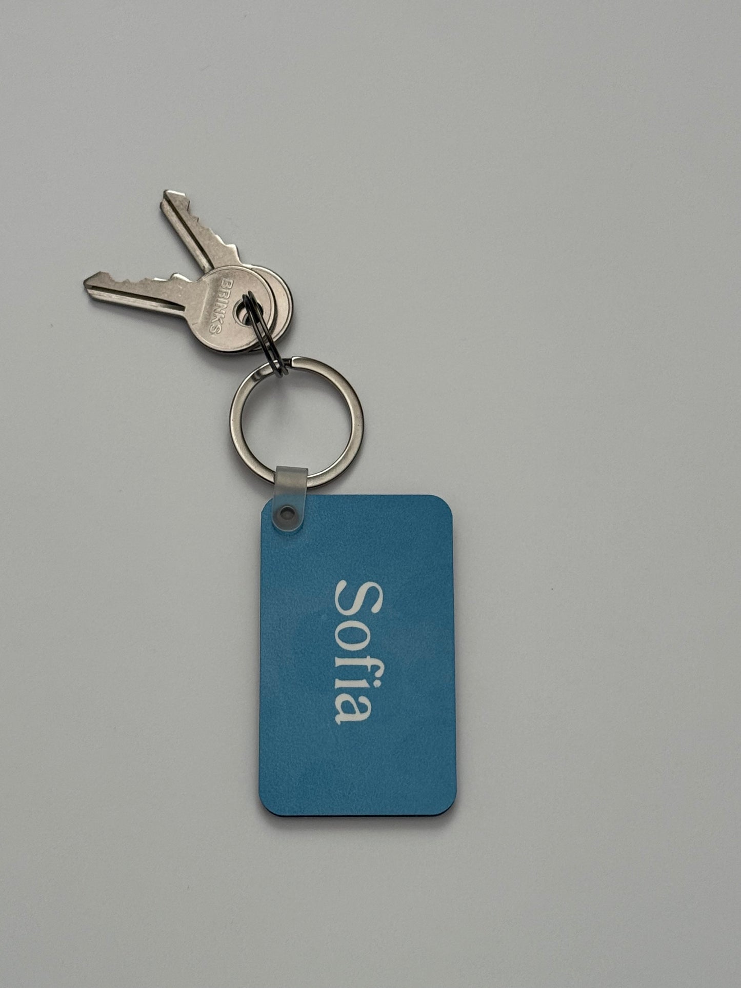 A personalized blue metal keychain with the name 'Sofia' in white, attached to a silver split keyring with two keys, on a neutral gray background. Roxzell