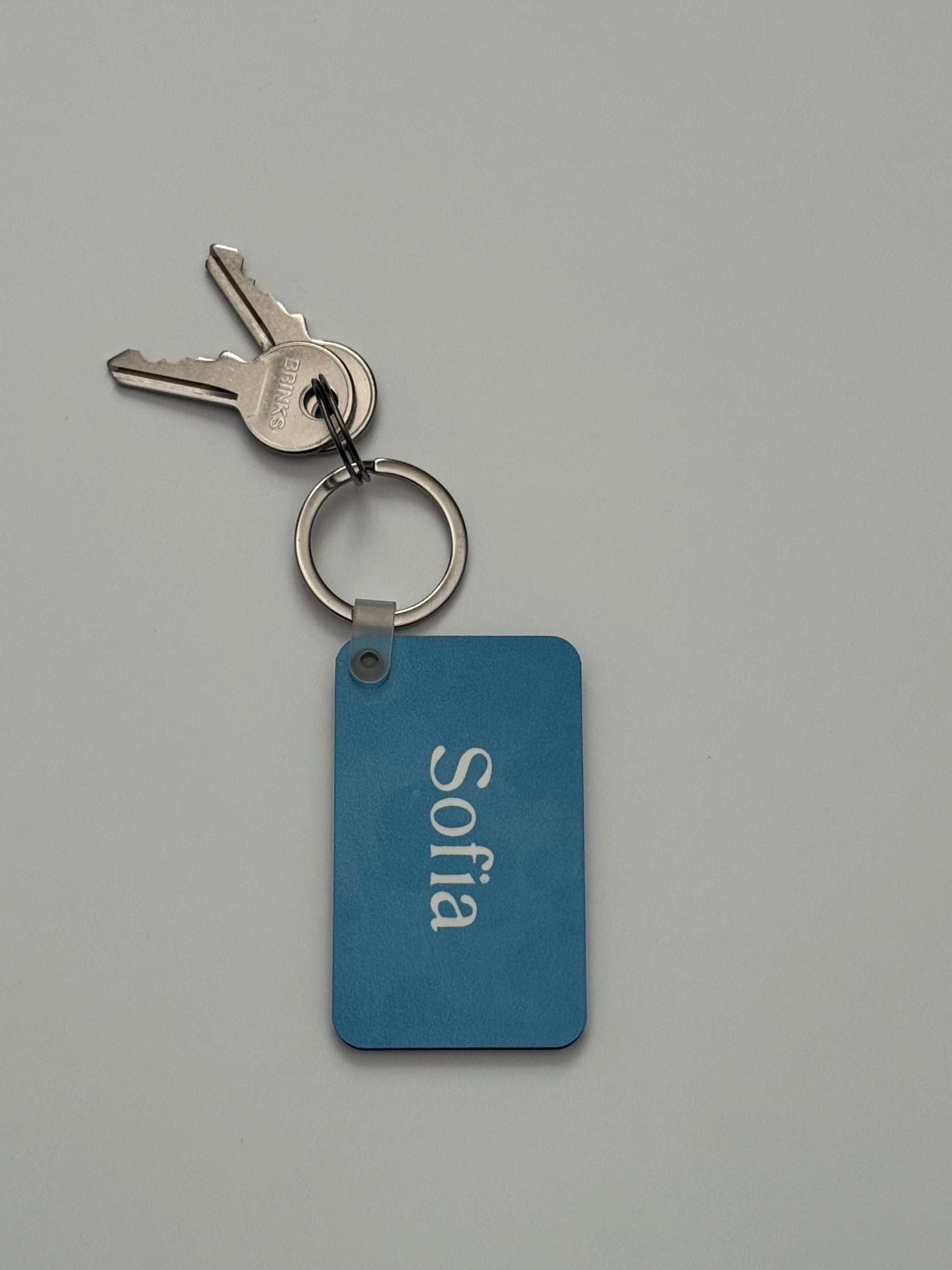 A personalized blue metal keychain with the name 'Sofia' in white, attached to a silver split keyring with two keys, on a neutral gray background. Roxzell