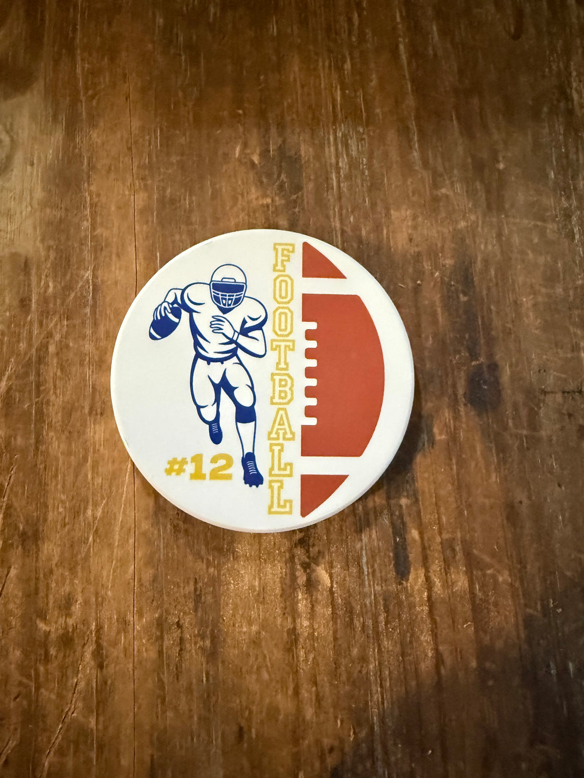 Round white coaster featuring a brown football graphic on the side, the word “FOOTBALL” in yellow letters, and a blue football player illustration wearing a helmet and uniform with “#12” printed in yellow, placed on a rustic wooden surface. Roxzell