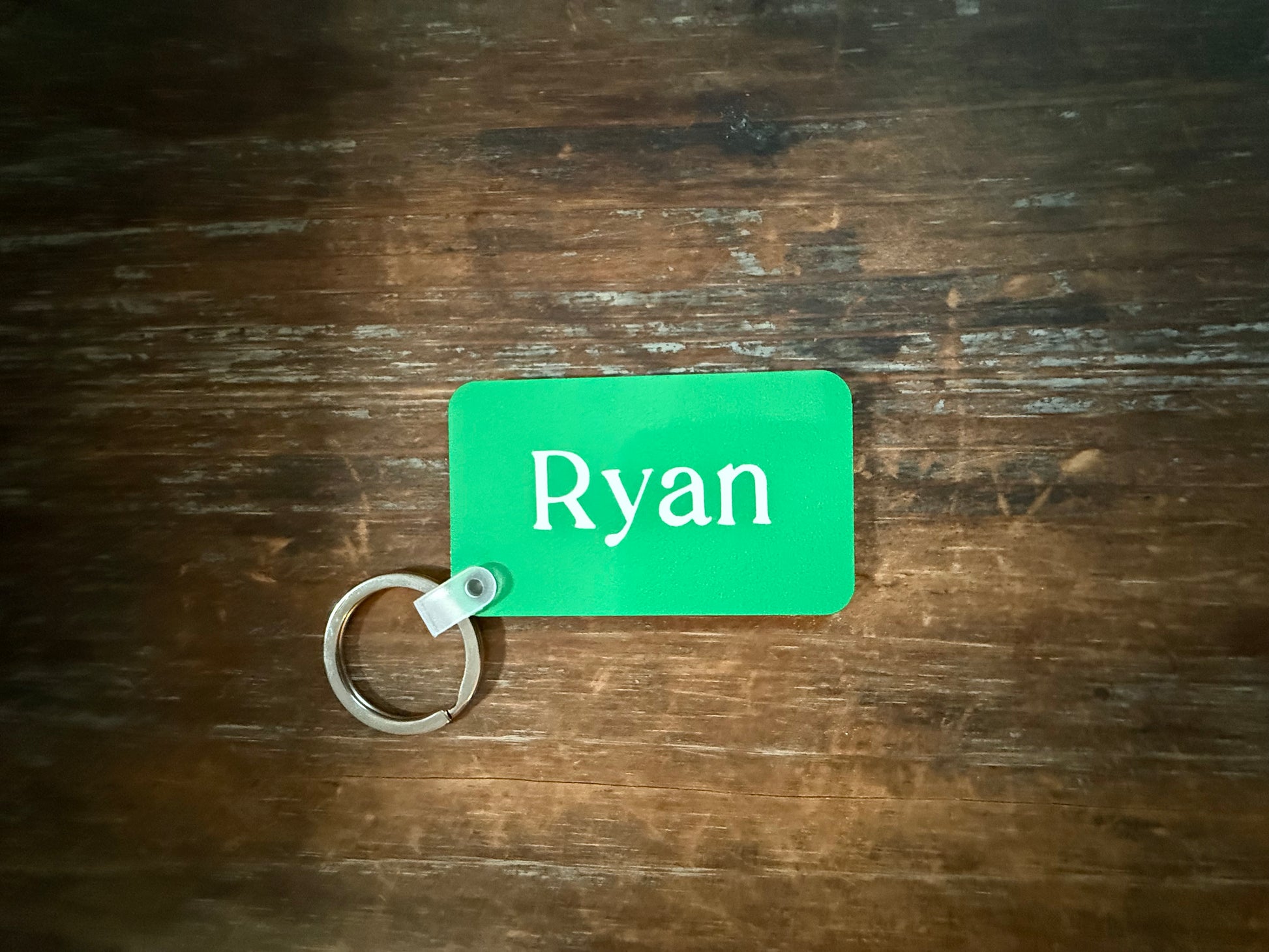 Green rectangular keychain with white text “Ryan” printed vertically, attached to a silver ring on a wooden surface. Roxzell