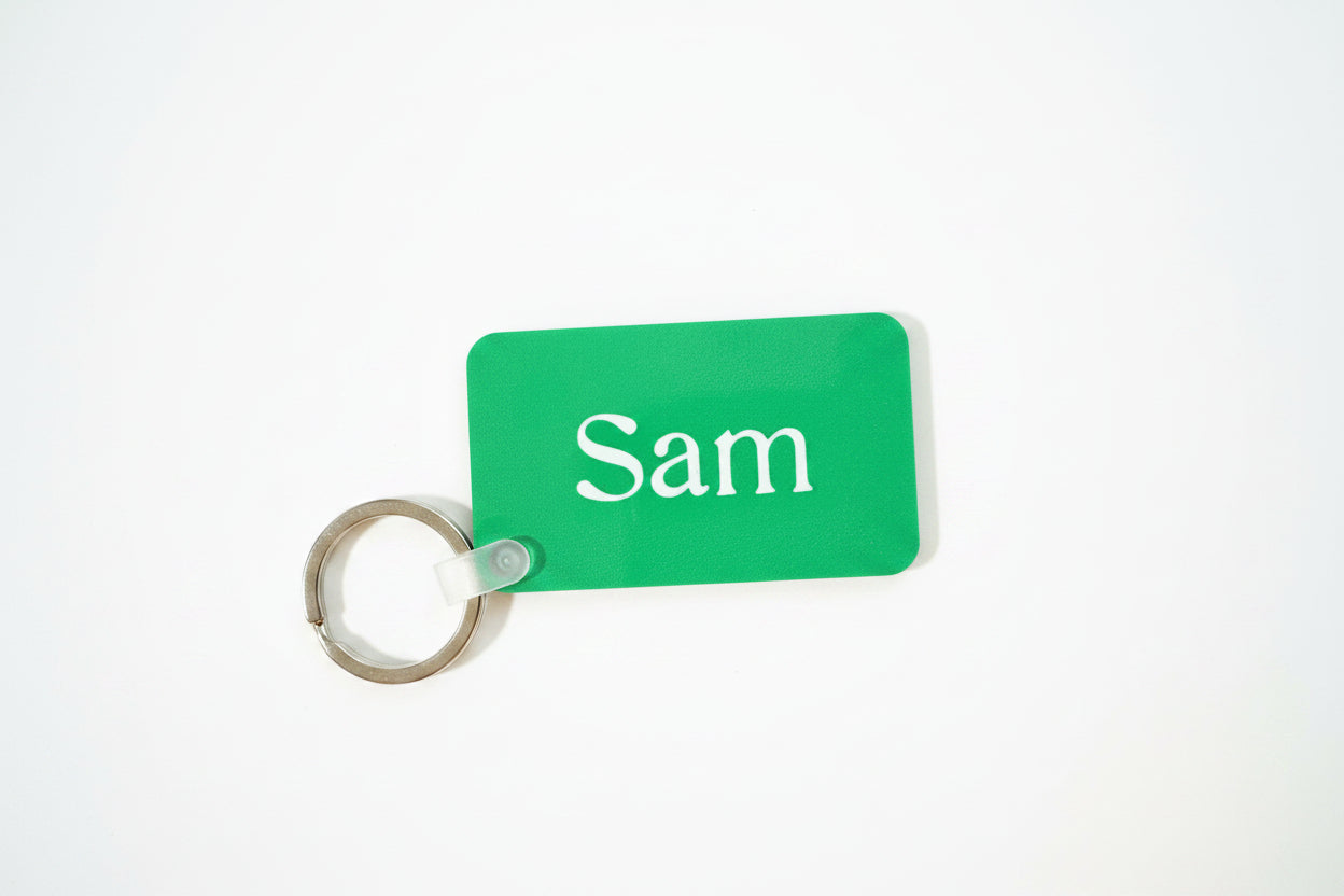 Green rectangular keychain with white text “Sam” on wooden background. Roxzell